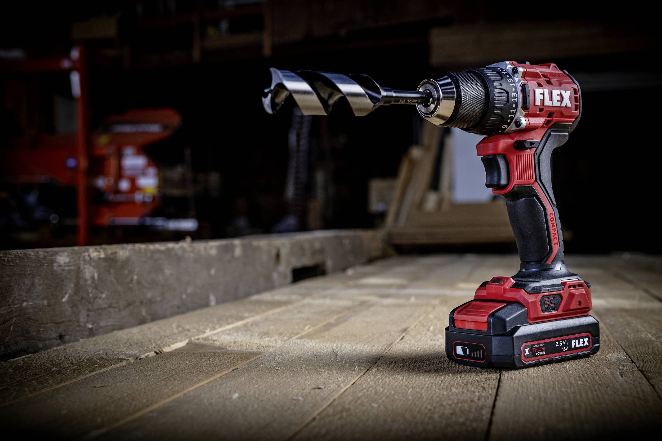 Cordless drill with a large spiral drill bit on a wooden workbench in a workshop. Distinctive with its red design and FLEX logo.