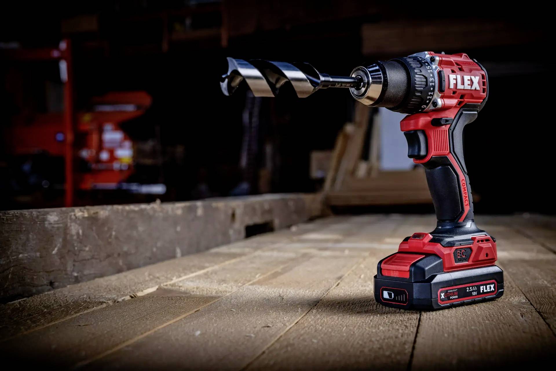 Cordless drill with a large spiral drill bit resting on a wooden board, in a workshop environment.