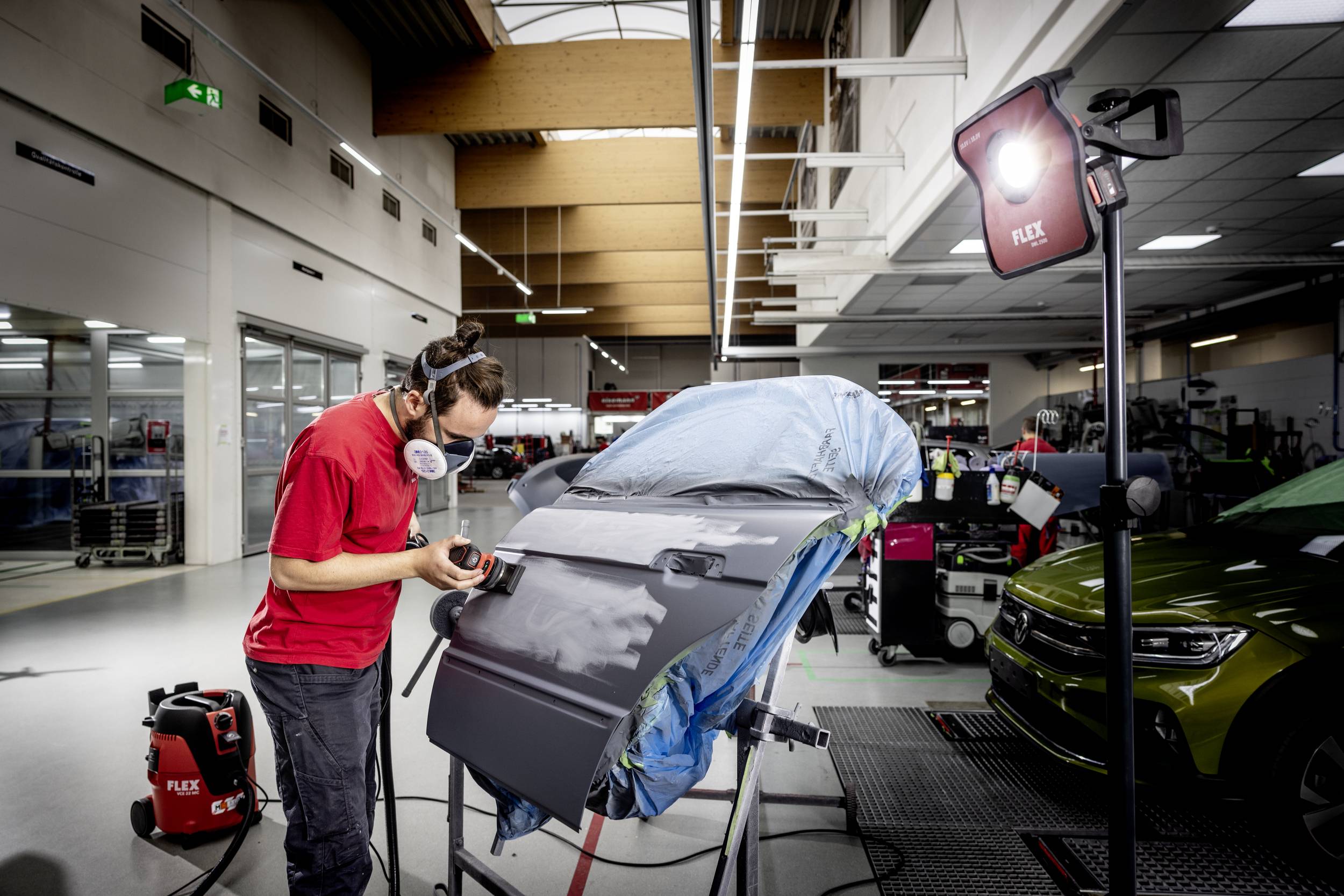 A man wearing a protective mask is sanding down a car bonnet in a workshop. In the background, other vehicles are parked.
