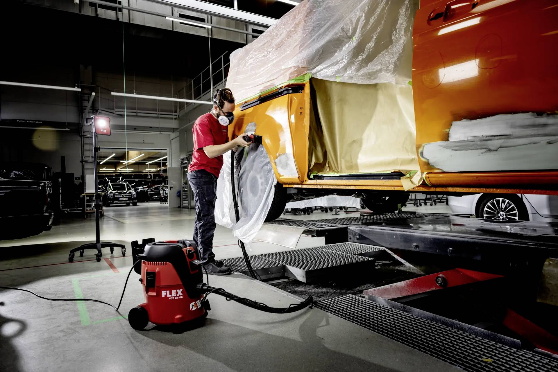 A person wearing a respiratory mask is grinding an orange car door frame in a workshop. A vacuum cleaner is in the foreground.