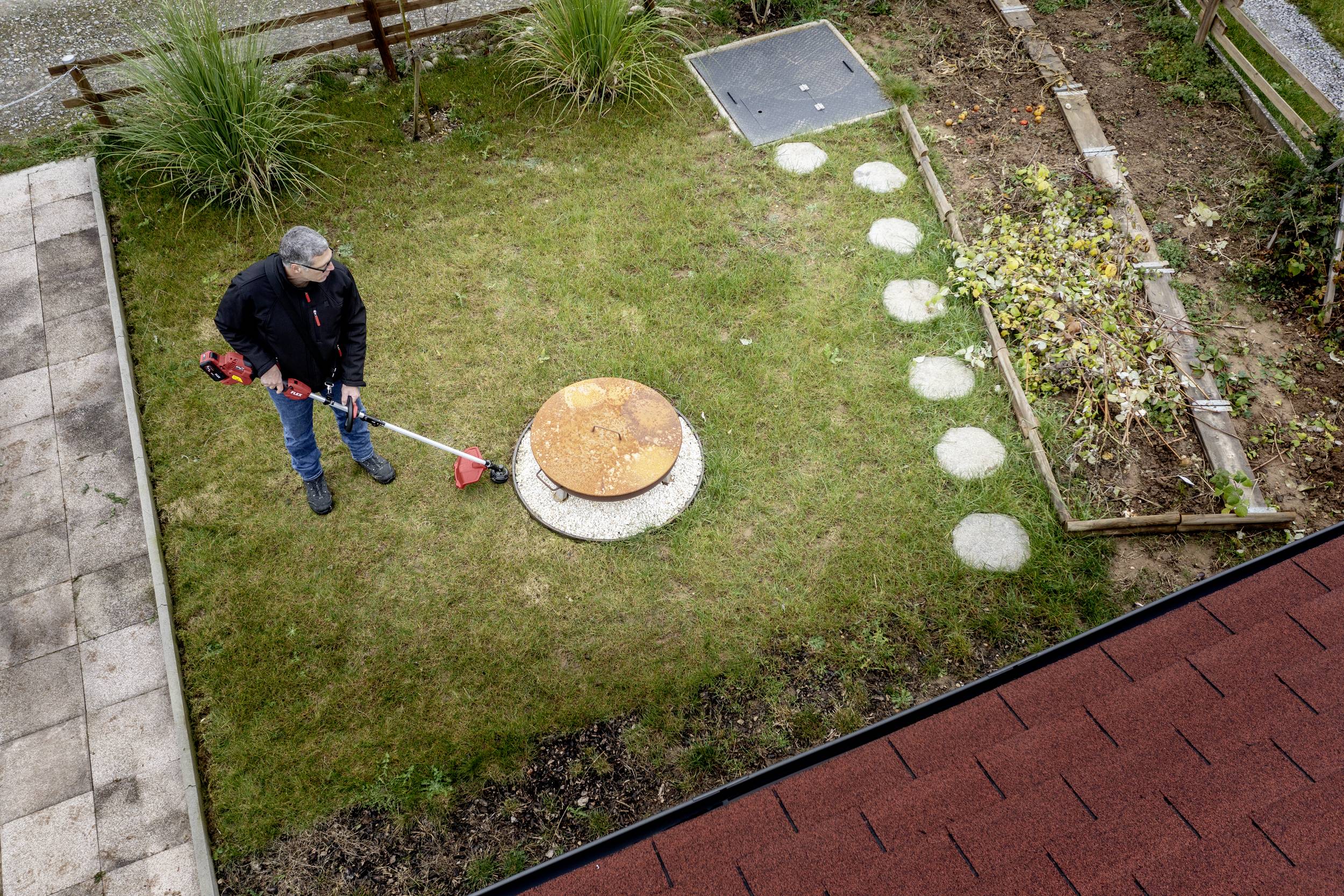 A man is mowing grass around a round manhole cover with a red lawn trimmer in the garden. Beside it are a small path and flower beds.
