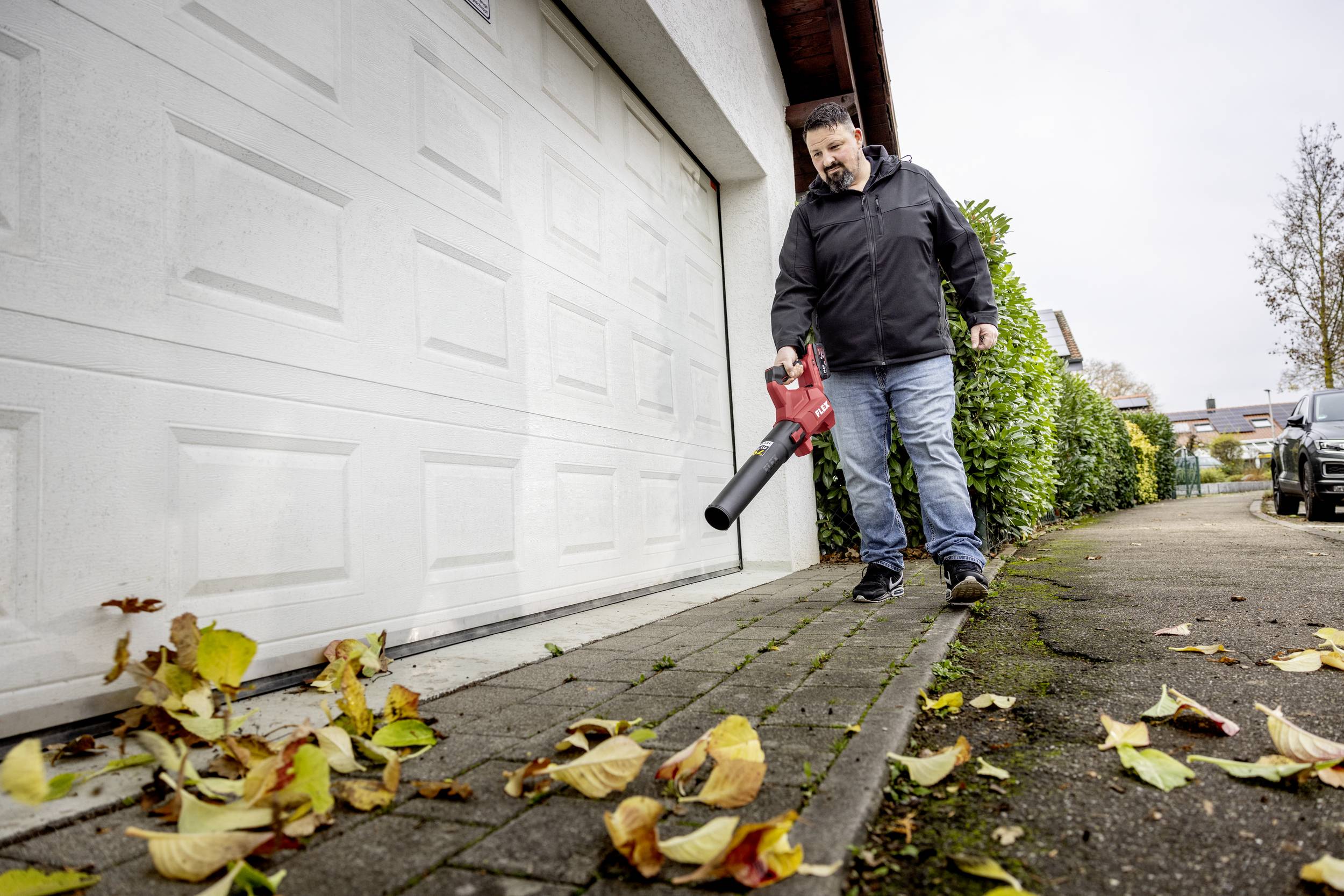 A person is blowing autumn leaves on a pavement with a leaf blower in front of a garage.