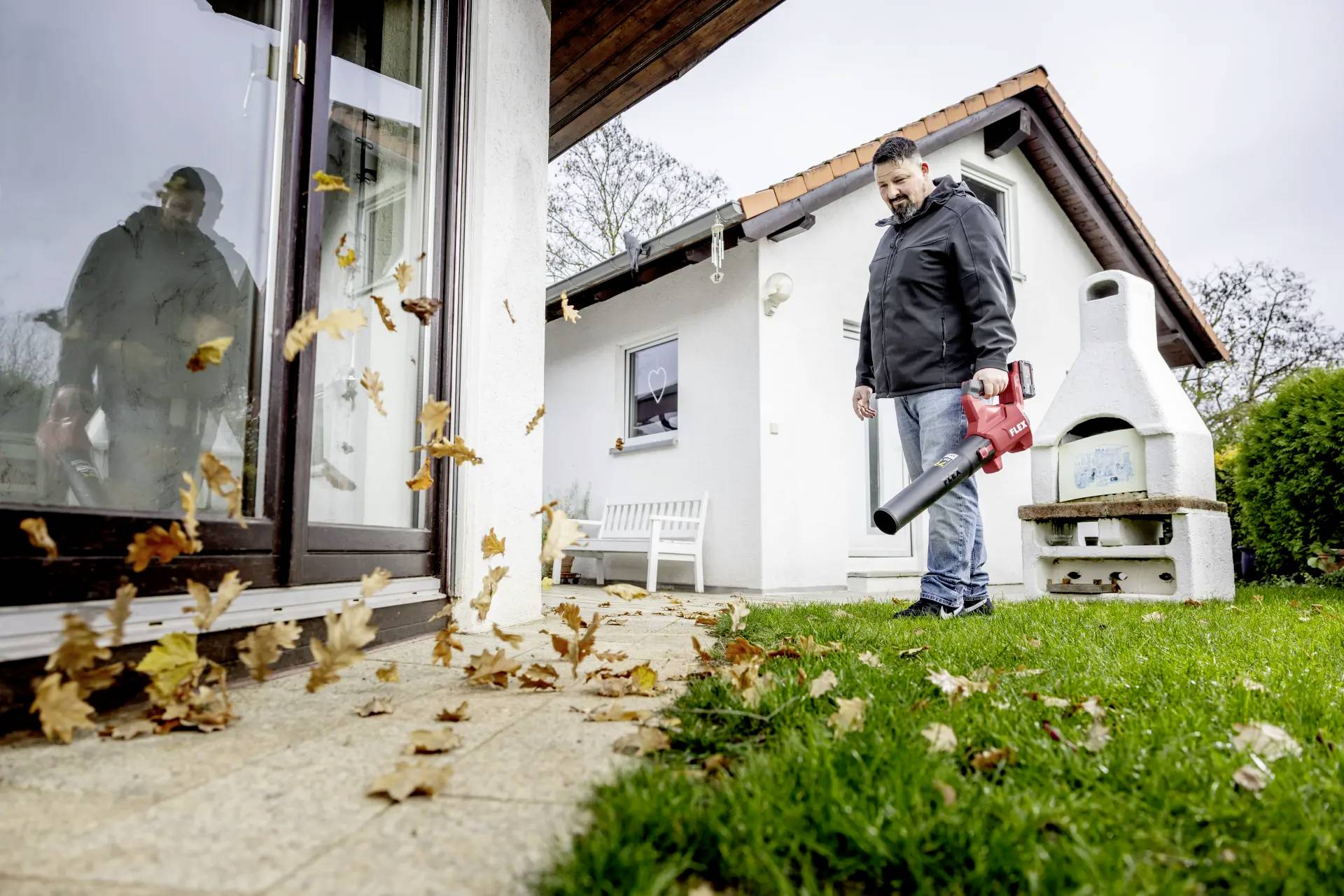 A person in the garden is using a leaf blower to remove leaves from the patio. A small white house stands in the background.