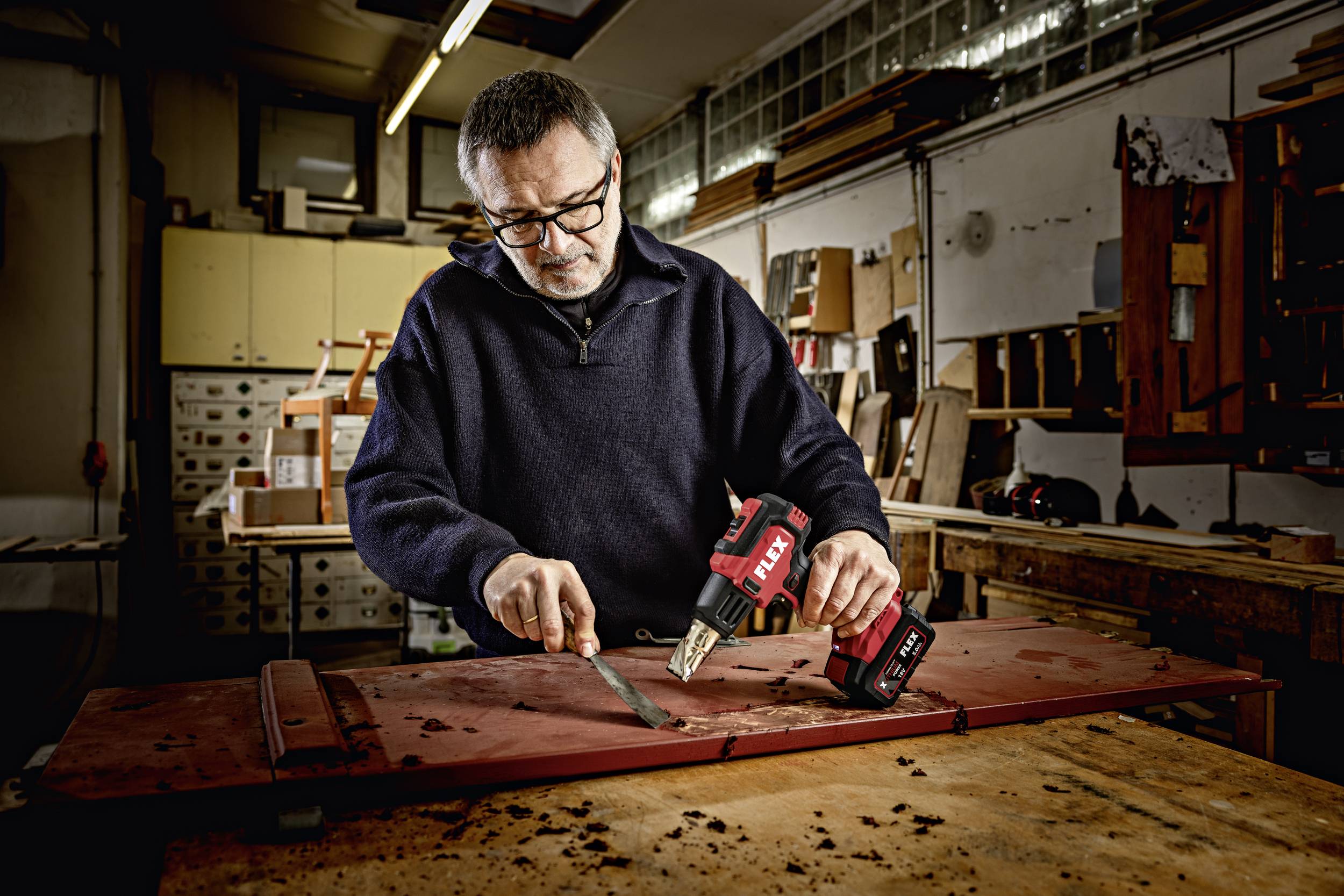 A man in a workshop is working on a wooden gate, using a putty knife and a cordless screwdriver.