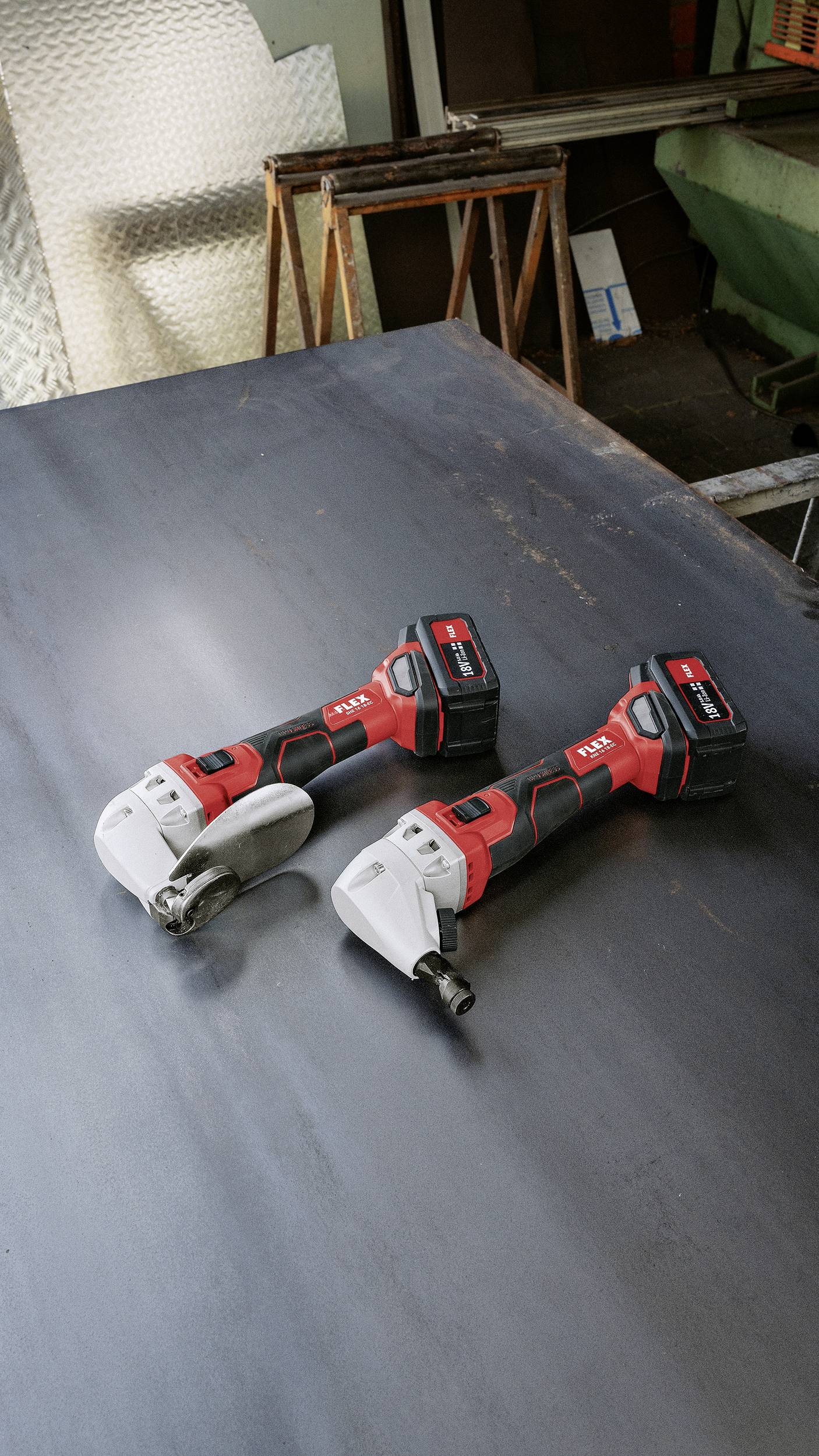 Two cordless angle grinders with red handles are lying on a metal table in a workshop.