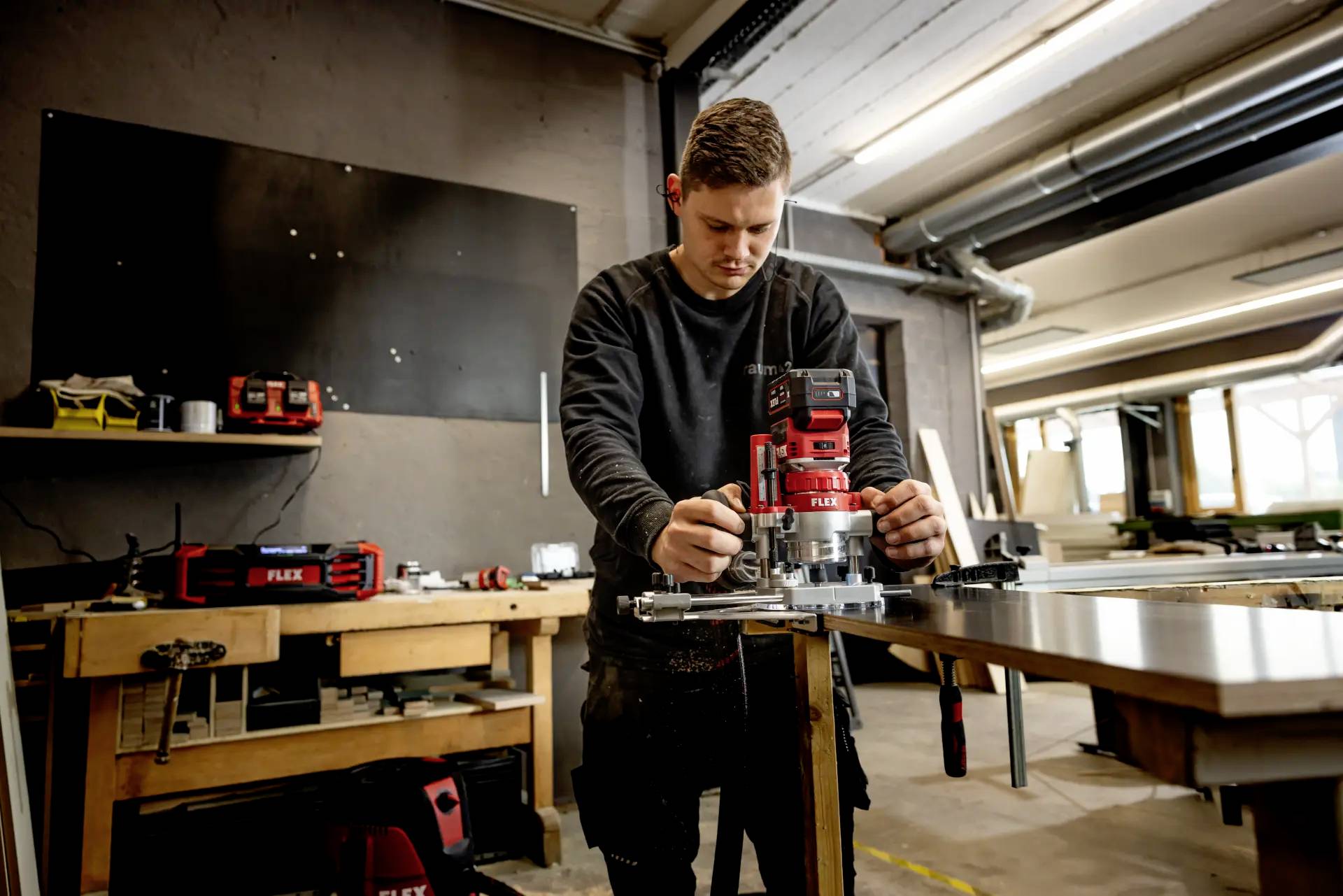 A person is working intently at a wood milling machine in a workshop. Various tools and work areas are visible in the background.