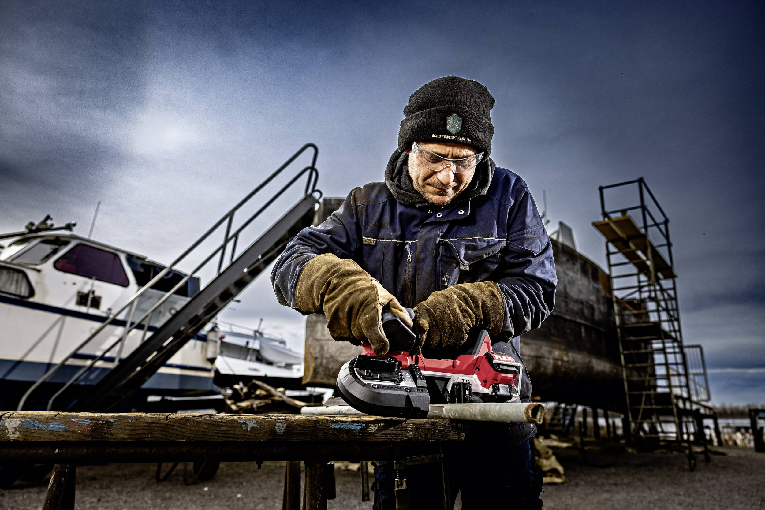 An older man wearing glasses and a cap is grinding a metal pipe with an angle grinder on a shipyard site. Boats are visible in the background.