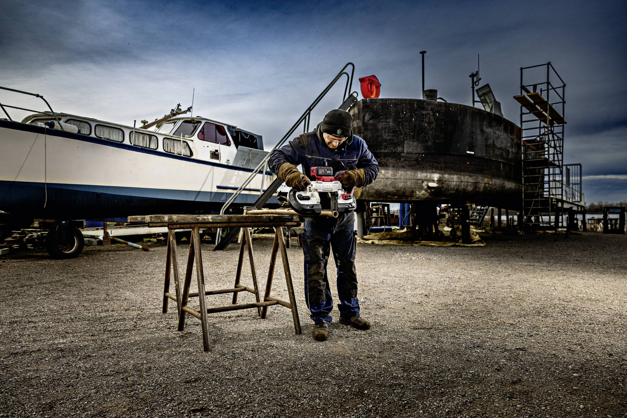 A worker in protective clothing is using an electric tool on a shipyard site, surrounded by boats and equipment outdoors.