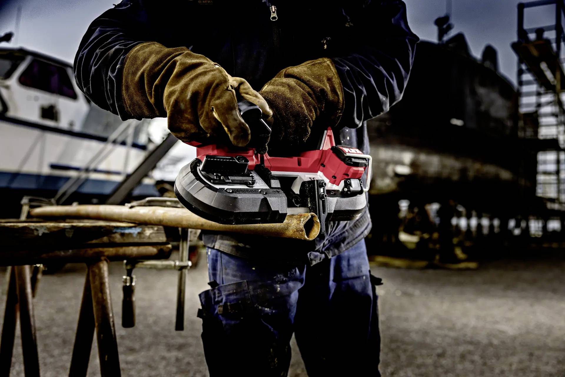 A worker wearing gloves is cutting a pipe with a battery-powered belt sander outdoors in a shipyard area.