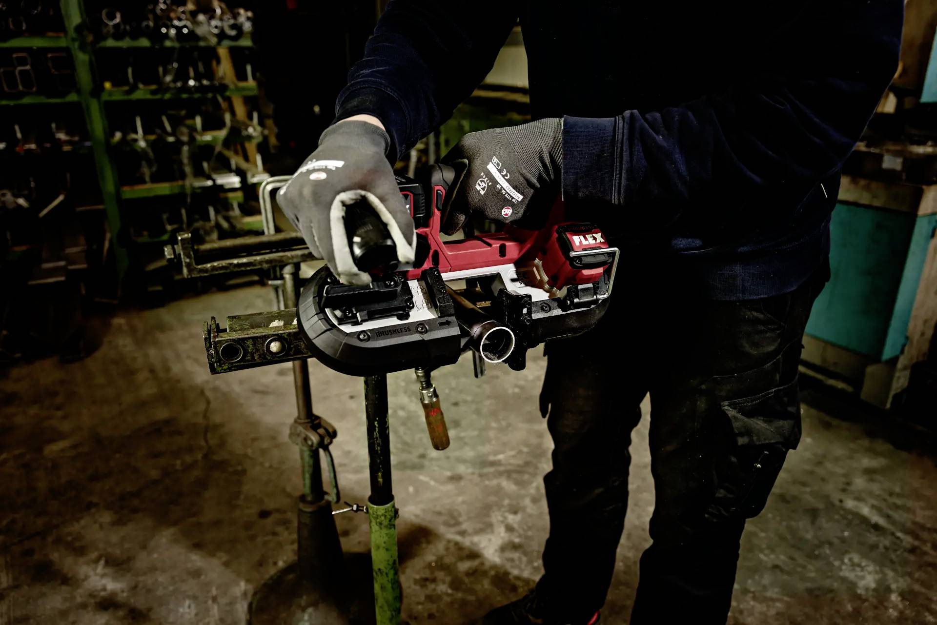 A person wearing dark work clothing and gloves is using a bandsaw to cut a metal pipe in a workshop.