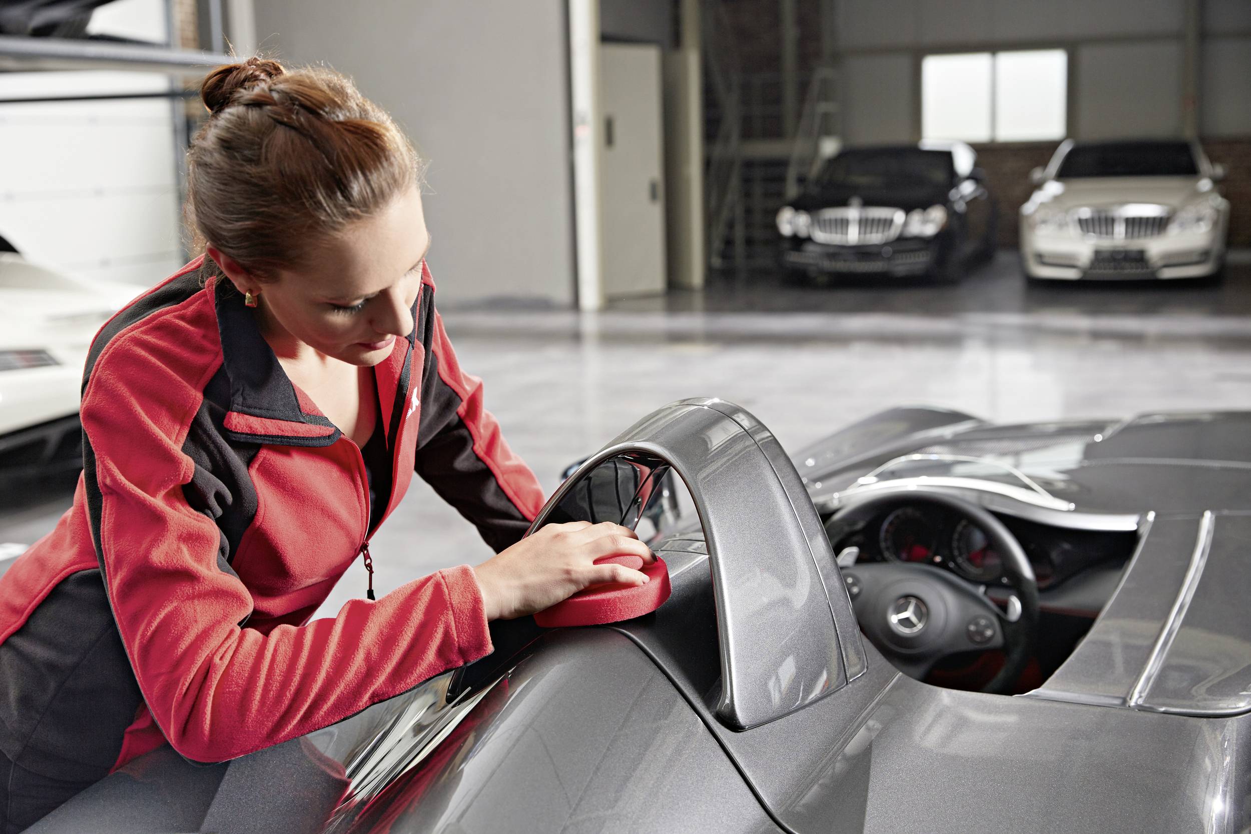 A woman is polishing a grey sports car in a car workshop. In the background, other luxury vehicles are parked.