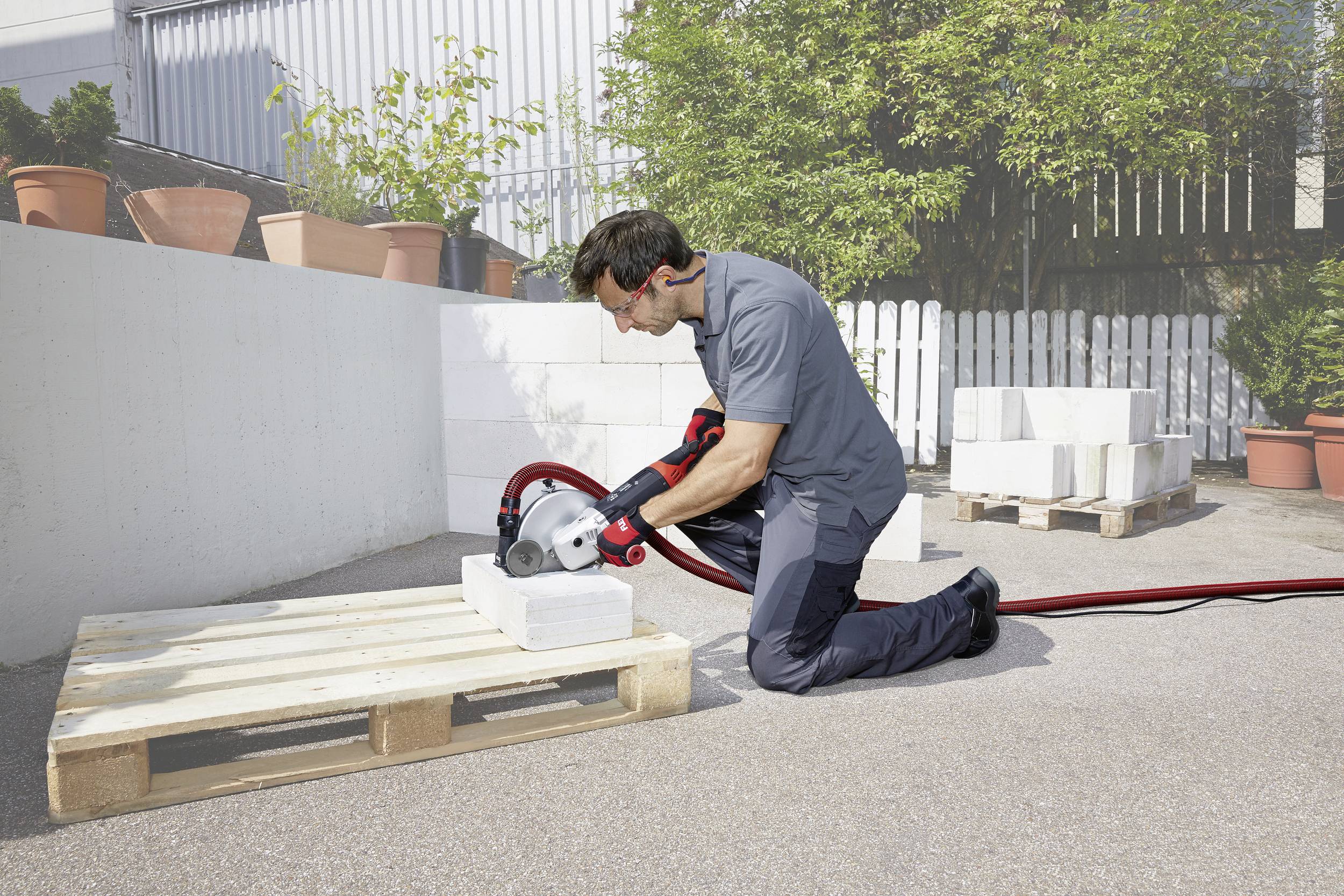 A man is kneeling on the ground, using a handheld tool to cut a stone block on a pallet outdoors.