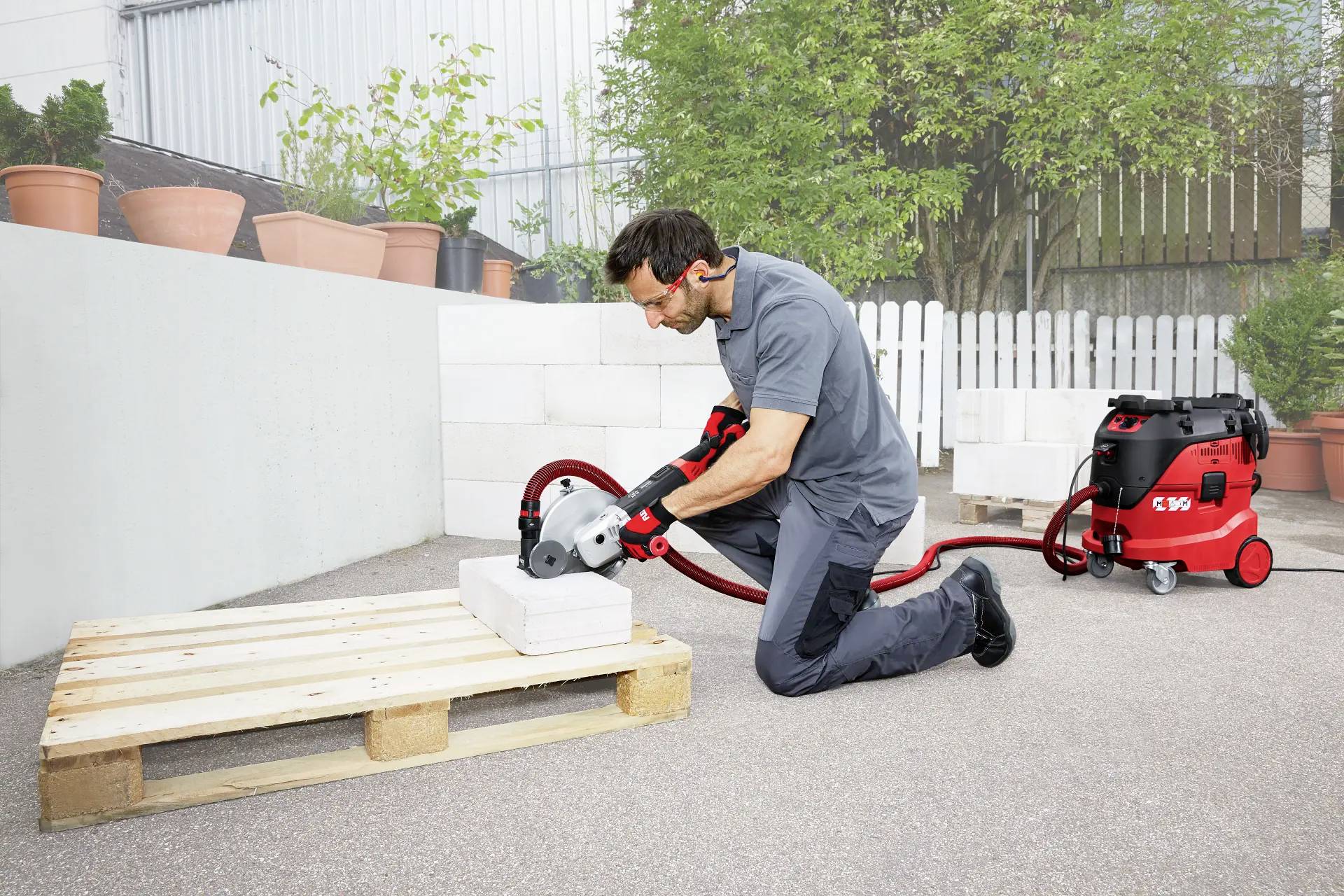 A man is kneeling and cutting a concrete block with an electric saw, connected to a vacuum cleaner. Outdoors, with plants in the background.