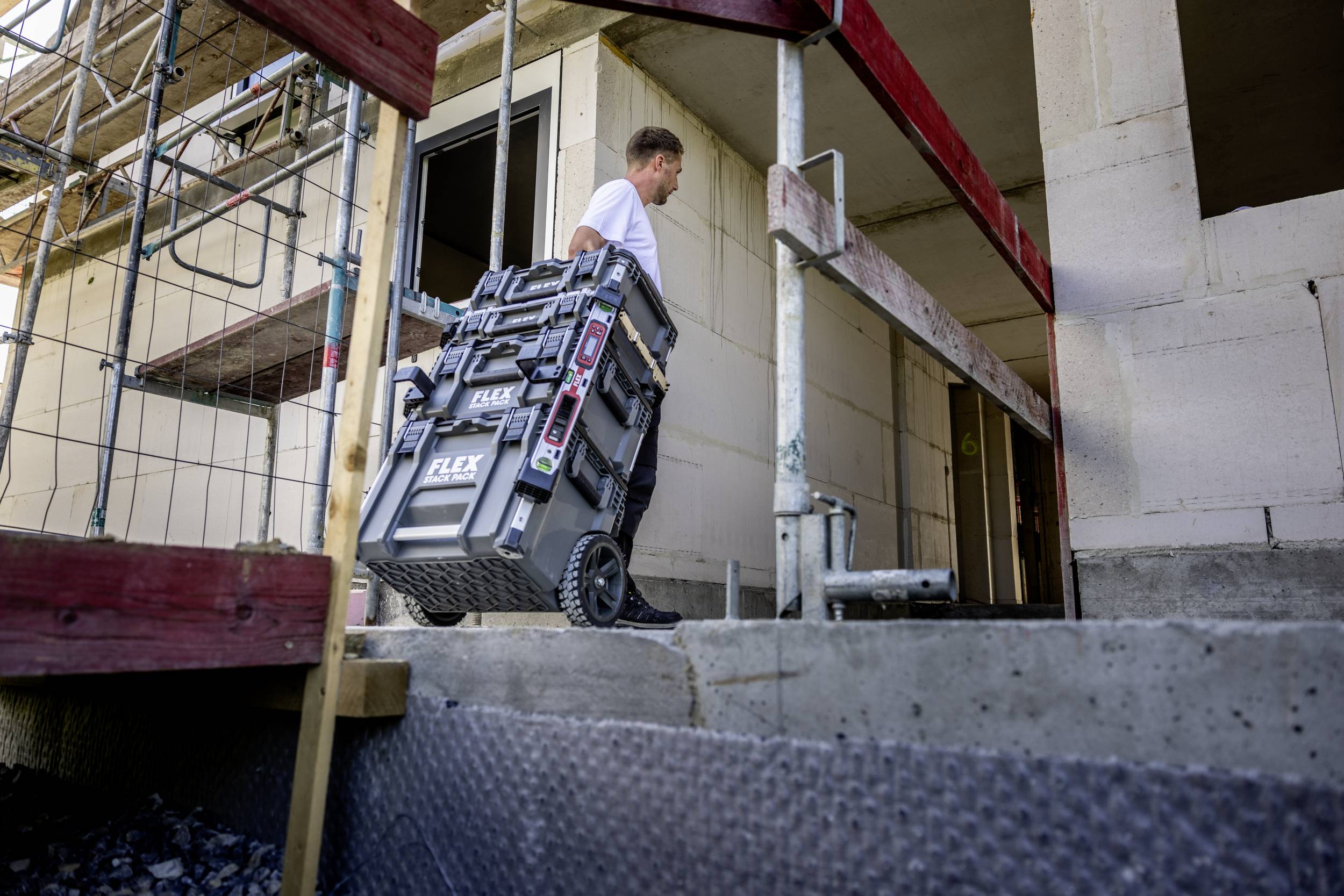 A construction worker pulls toolboxes on a trolley into the unfinished shell of a building. Scaffolding can be seen in the background.