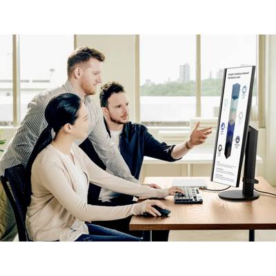 Three people are examining a large computer monitor diagram in the office. The man is pointing at the diagram while the others listen attentively.