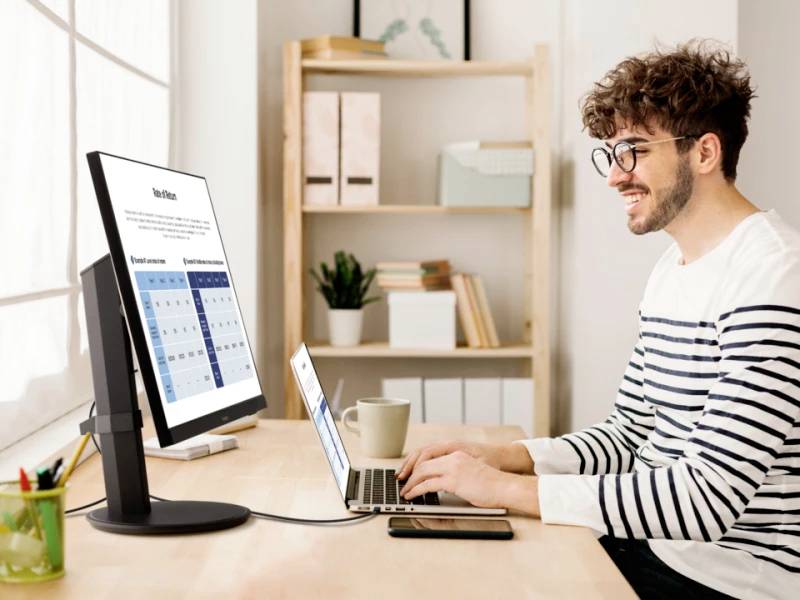 A man is working on a laptop connected to a large monitor. He is sitting at a desk with shelves in the background.