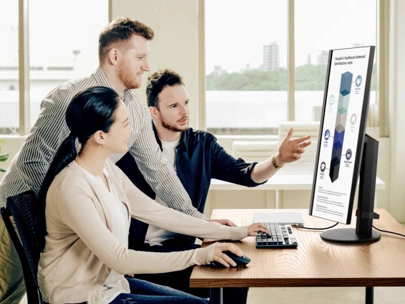 Three people are working together at a computer screen displaying a diagram. One person is pointing at the screen. Office setting in the background.