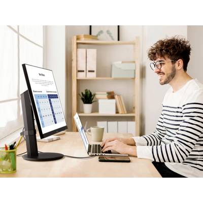 A man is working on a laptop at a desk, with the monitor displaying a diagram. Shelves with folders and decorations are visible in the background.