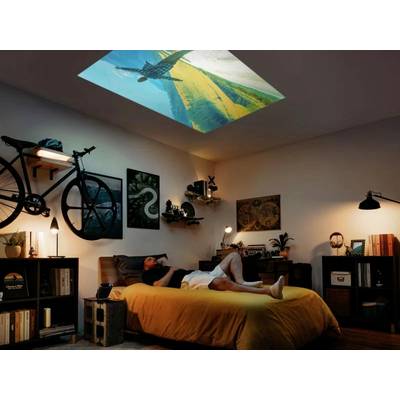 A man is lying relaxed on a bed in a cosily decorated room. A film is being projected onto the ceiling.