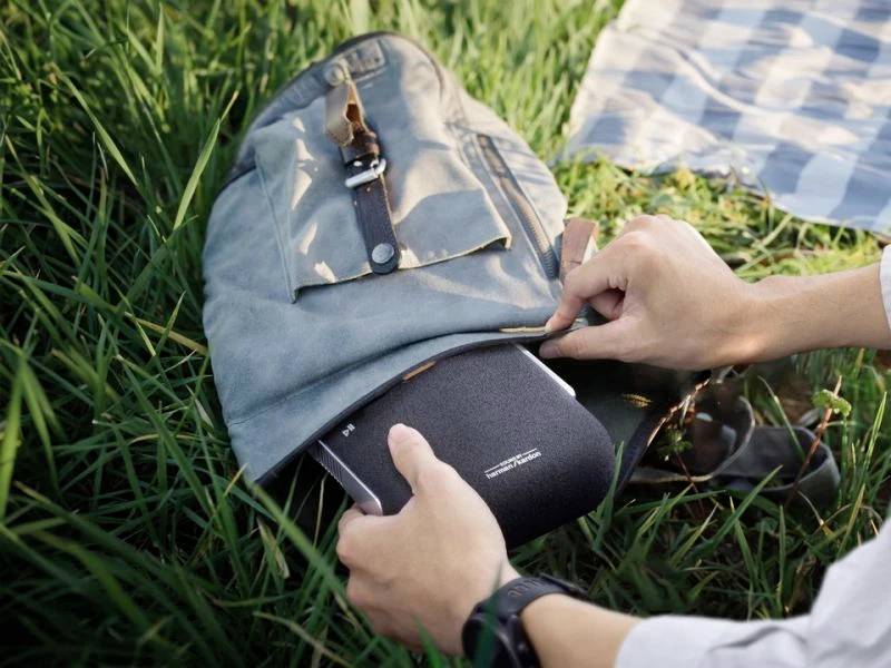 A person is stowing a black portable device in a blue rucksack lying on the grass. The surroundings appear relaxed and natural.