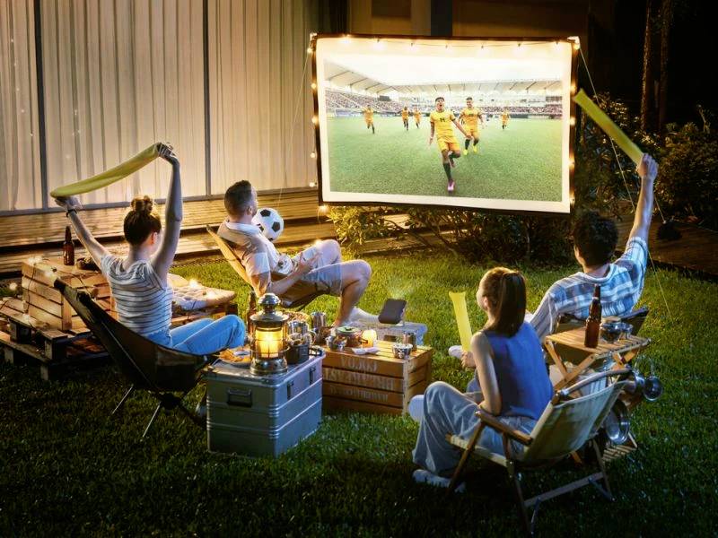 'Four people are sitting in the garden watching a football match on an outdoor screen. Drinks and snacks are at the ready.'