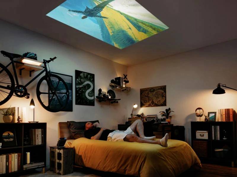 A man is lying on a bed in a cosily decorated room, gazing at an image projected onto the ceiling.