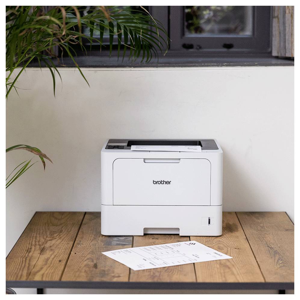 A white printer sits on a wooden table, with a printed sheet of paper beside it. In the background, a window and plants can be seen.