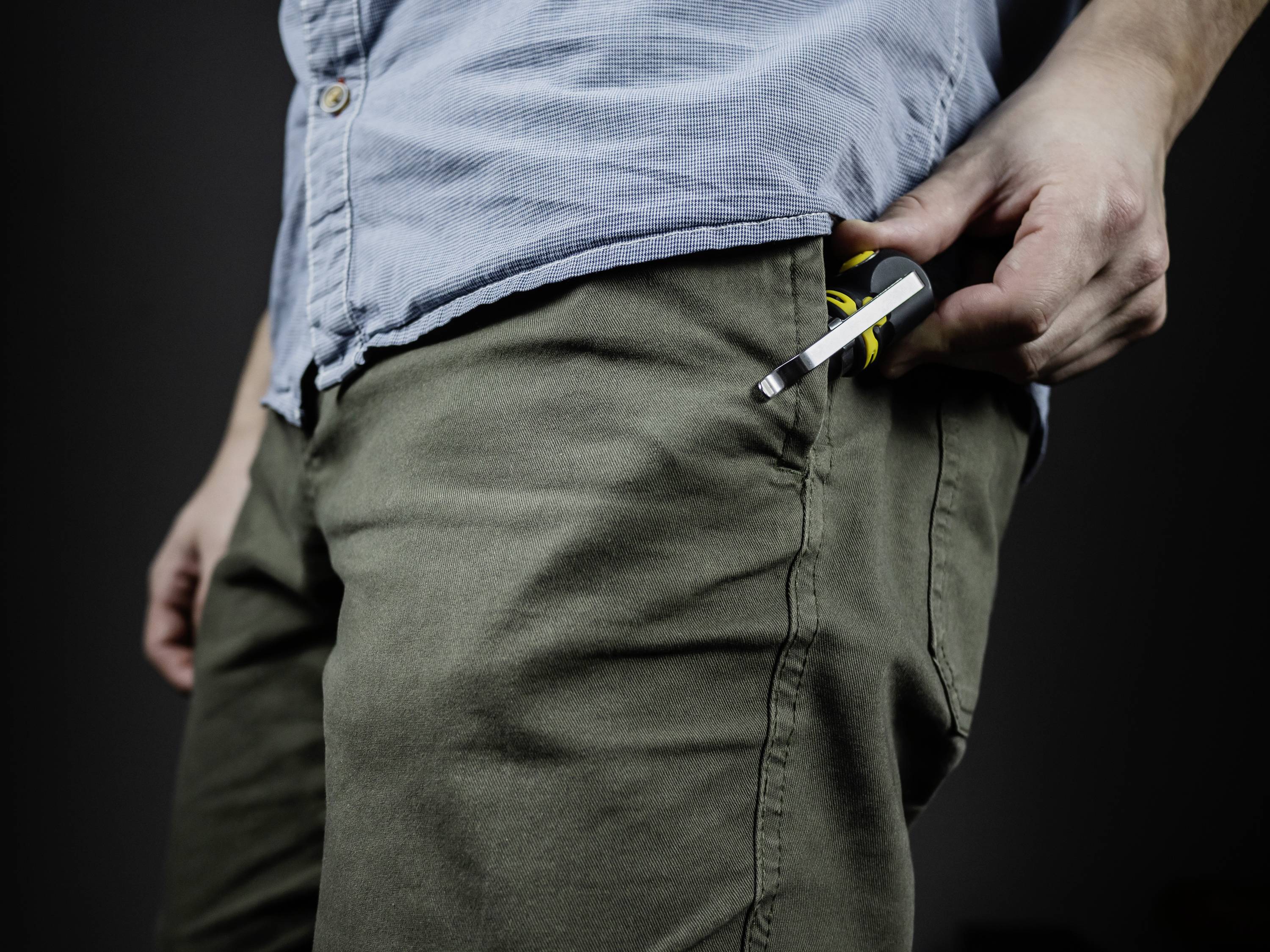 A man in a blue shirt and green trousers is putting a screwdriver into his trouser pocket.
