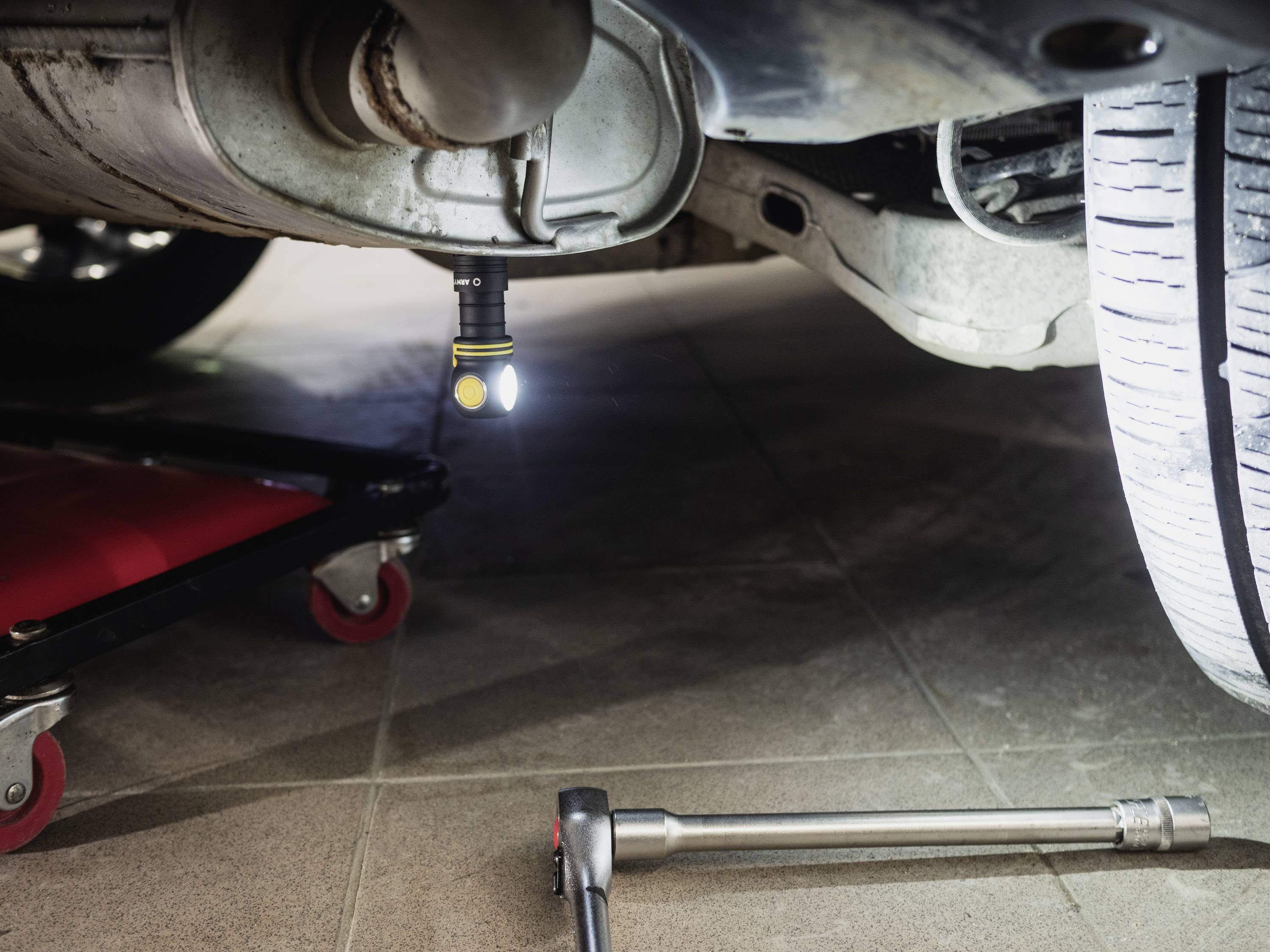 Car in workshop with torch under the chassis. A vehicle lift and spanners are visible, repair in progress.