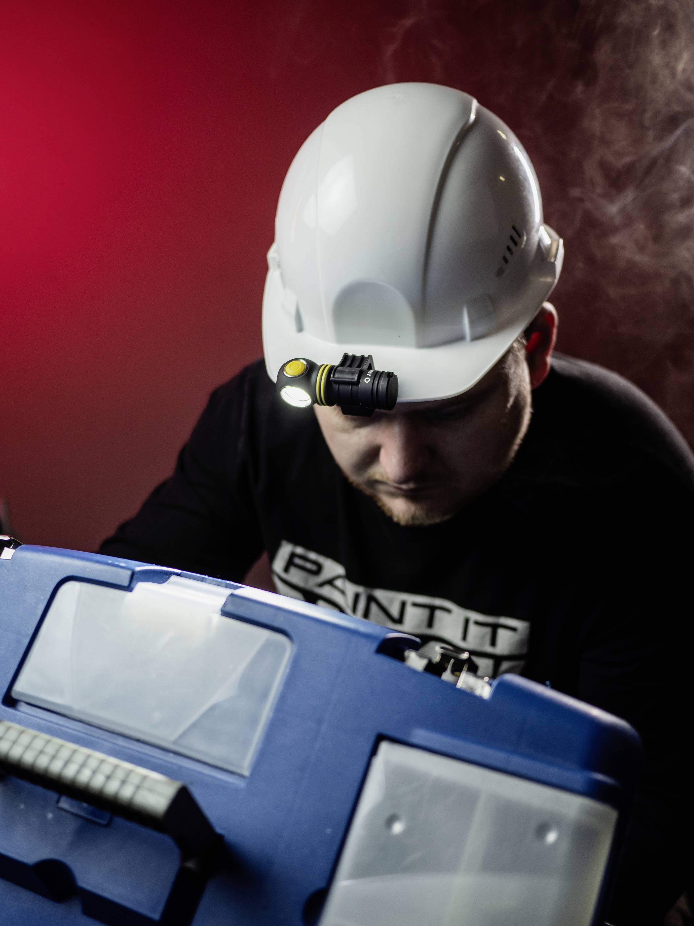 A construction worker with a white hard hat and head torch inspects a blue toolbox in a dimly lit room.