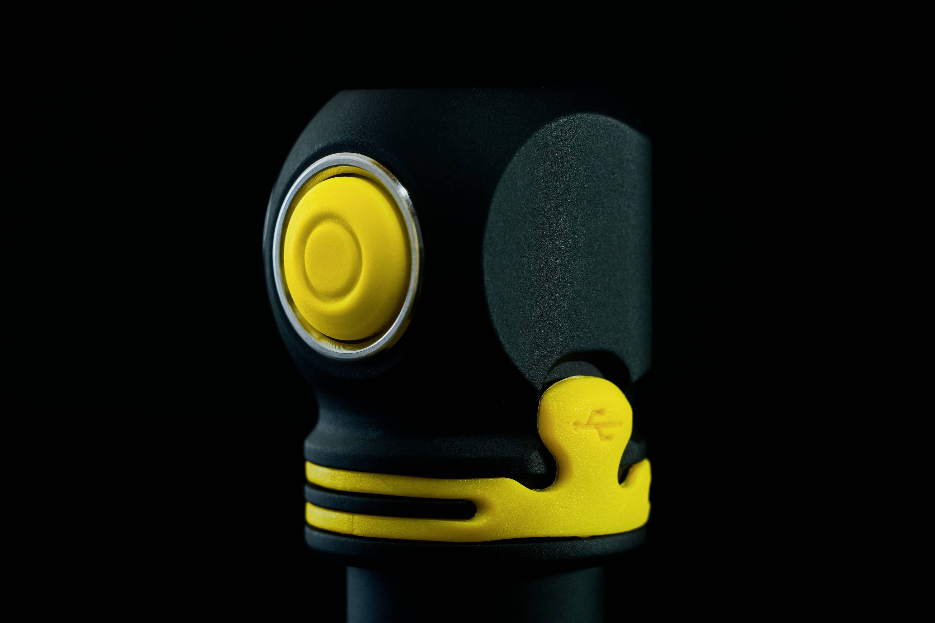 A black device with a yellow button against a black background. The button is round and surrounded by a silver ring.
