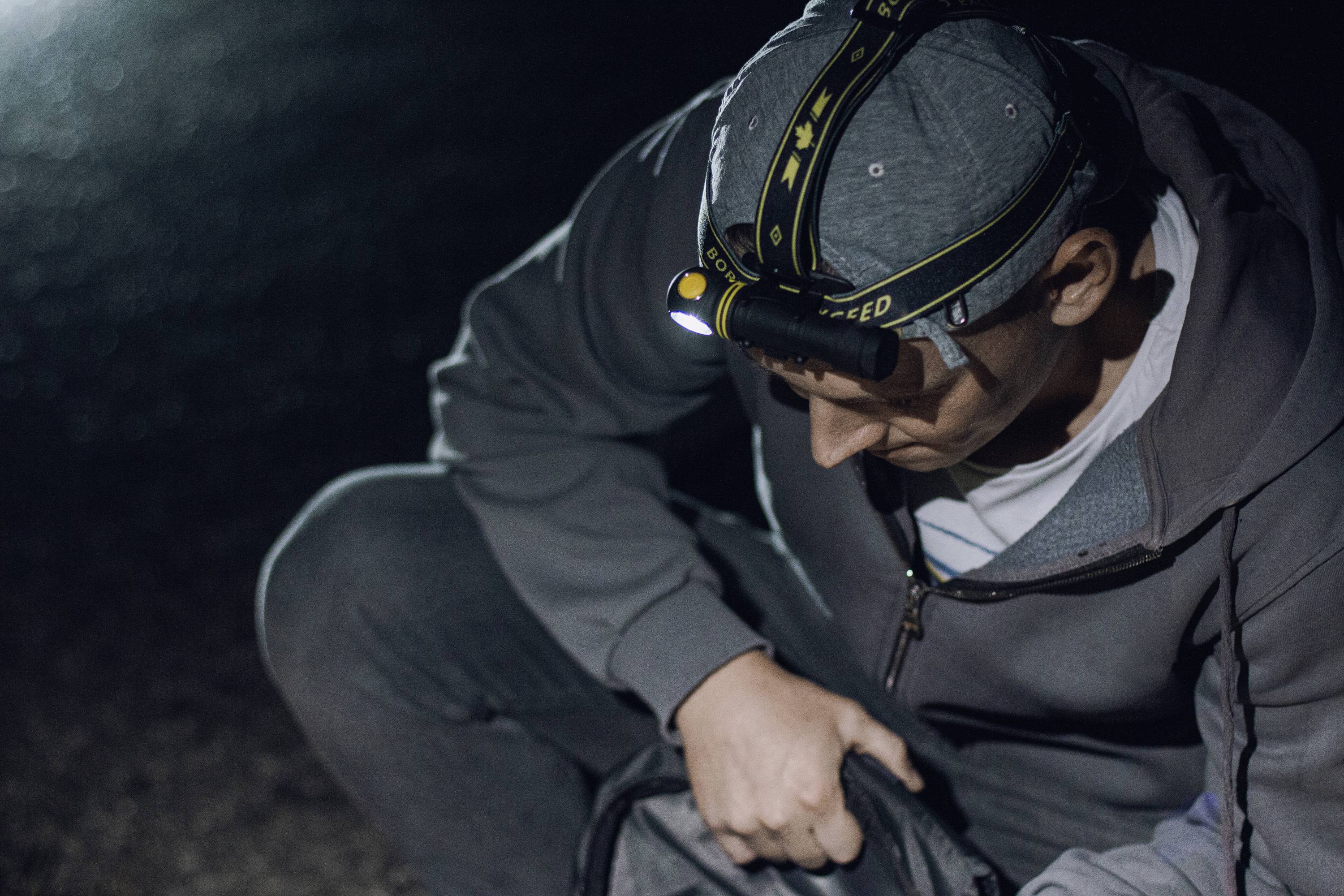 A person kneels outdoors at night, wearing a head torch while searching through a bag.