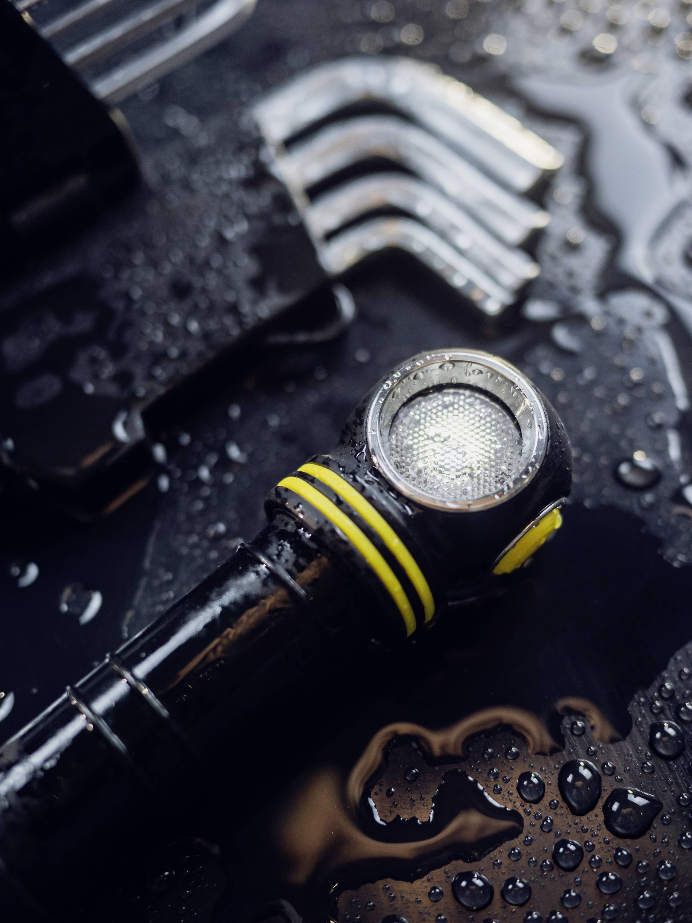 Torch with a yellow stripe lies on a wet, glossy surface. Water droplets emphasise a rainy or damp environment.<br><br>Note: I've used "torch" instead of "flashlight" to align with British English terminology, and used British spelling of "emphasise".