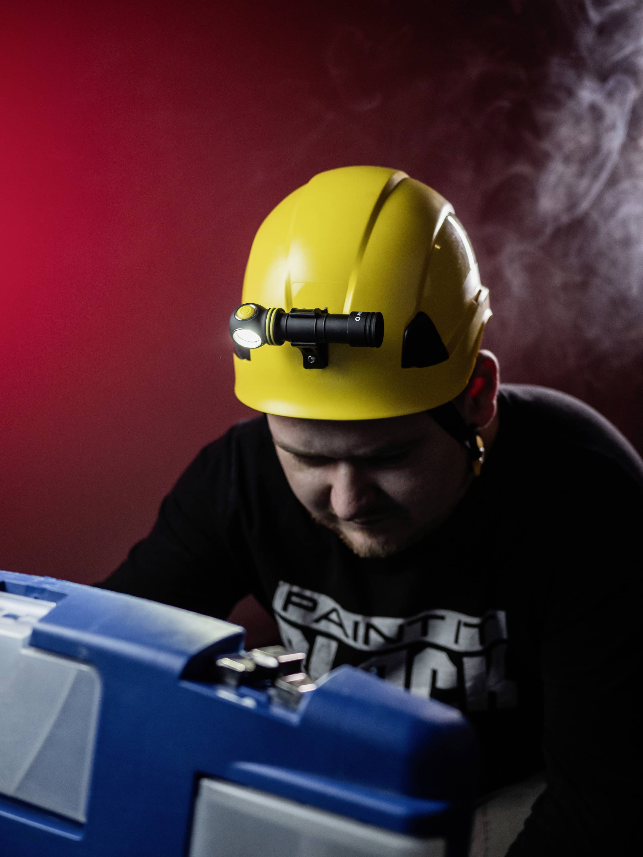 A man wearing a yellow safety helmet with a head torch, leans over a blue toolbox in the light of a red background lighting.