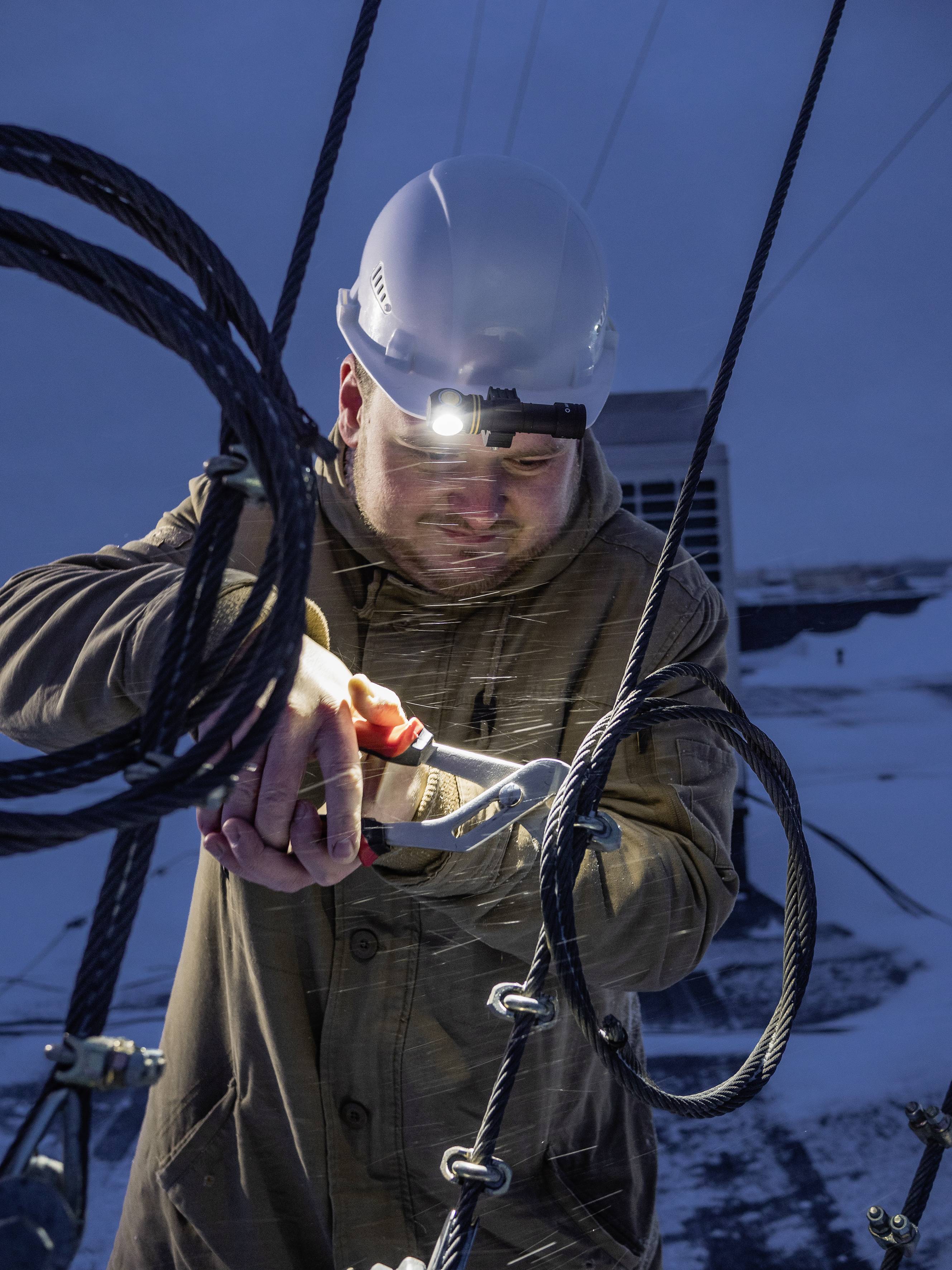 A worker wearing a hard hat and head torch repairs cables outdoors at dusk. The background shows snow and technical installations.