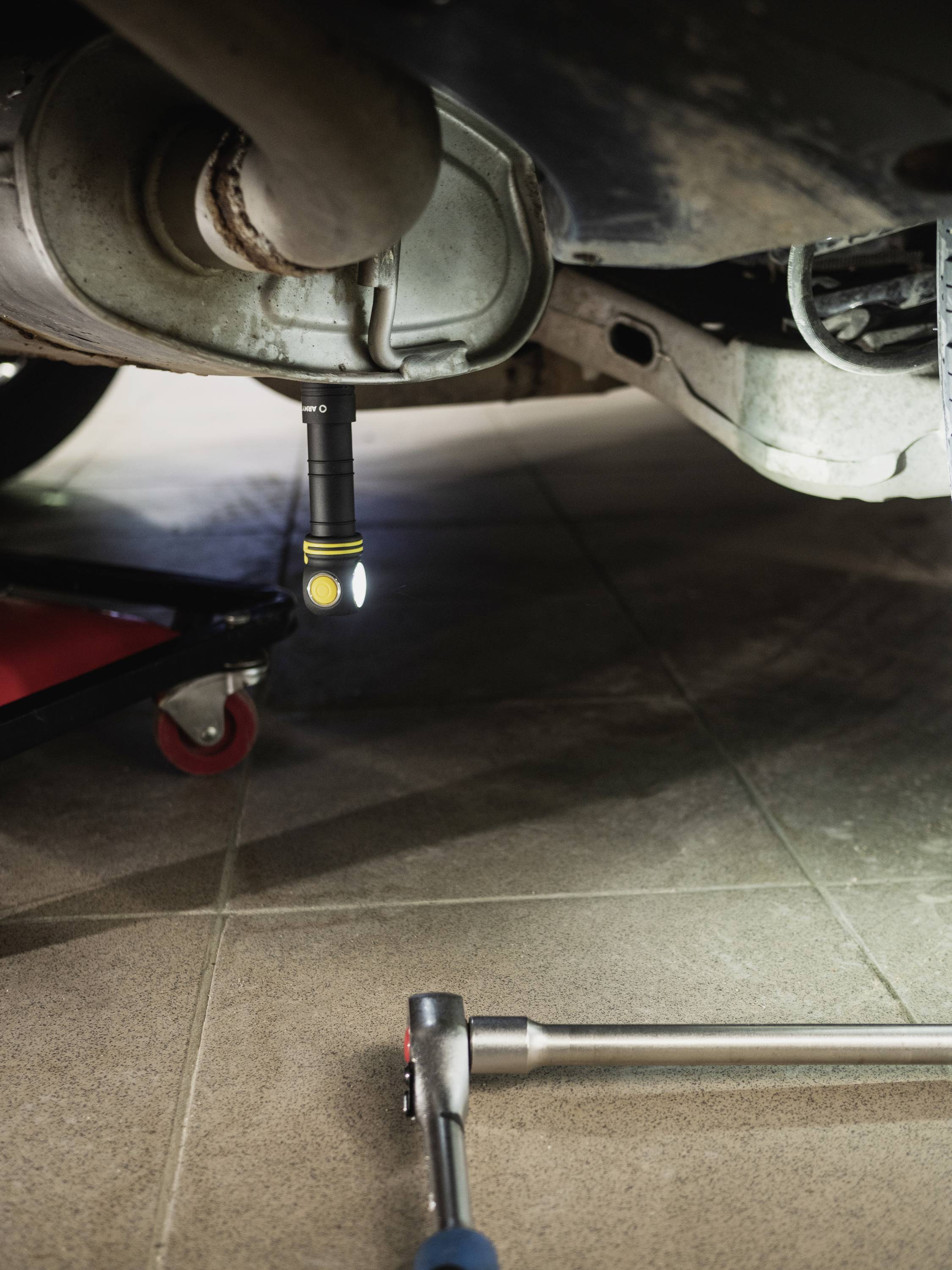 Exposed car exhaust system with a torch illuminating it underneath. A tool is lying ready on the ground.