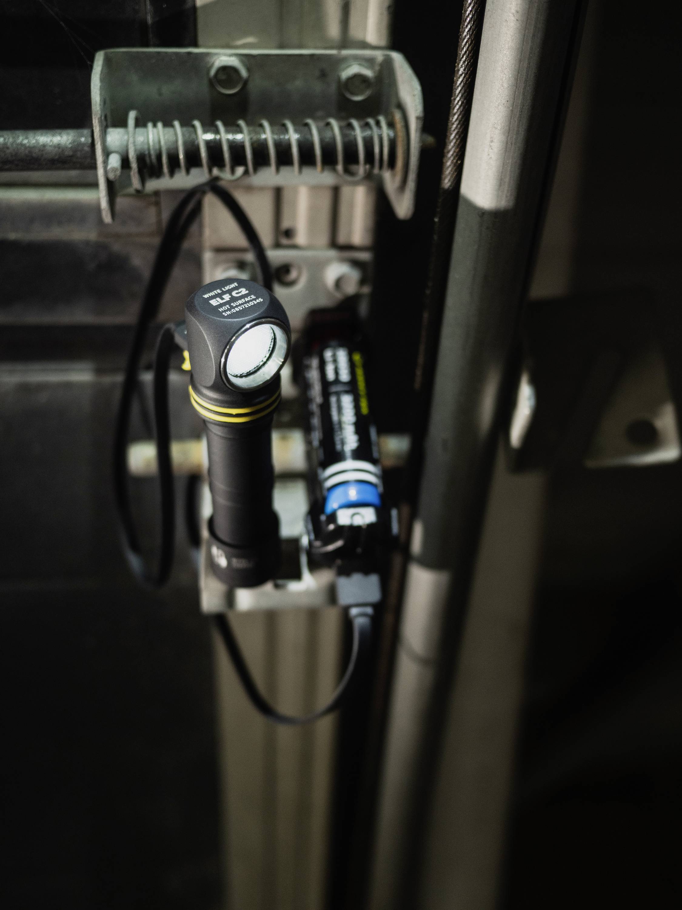 Torch with a circular LED and blue-black battery, mounted on a metal door next to a cable.