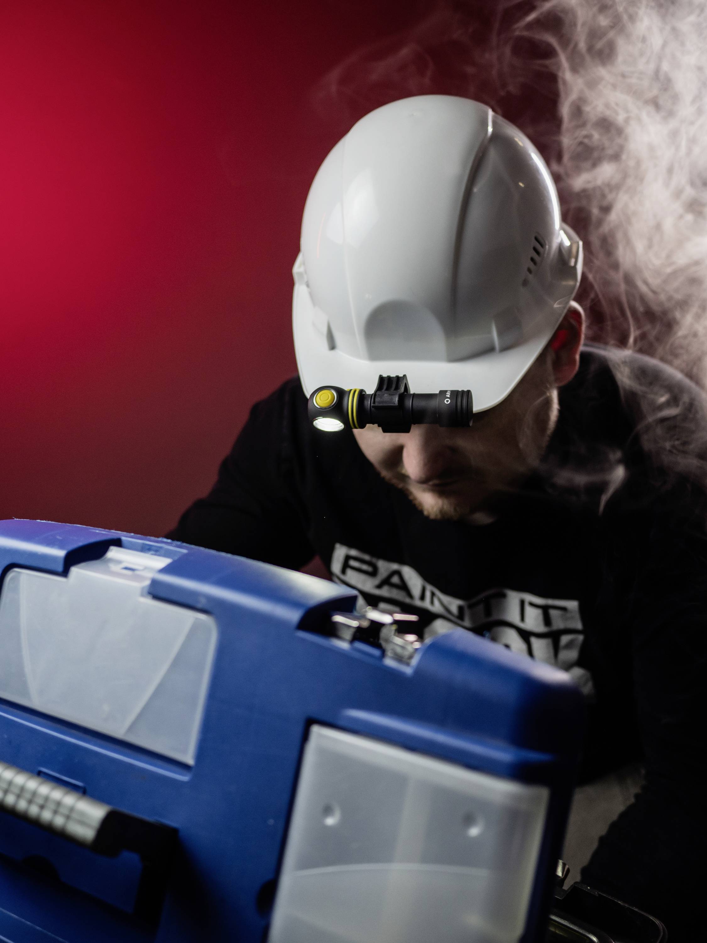 A person is wearing a protective helmet with a head torch and is working intently on a blue toolbox. Smoke in the background.