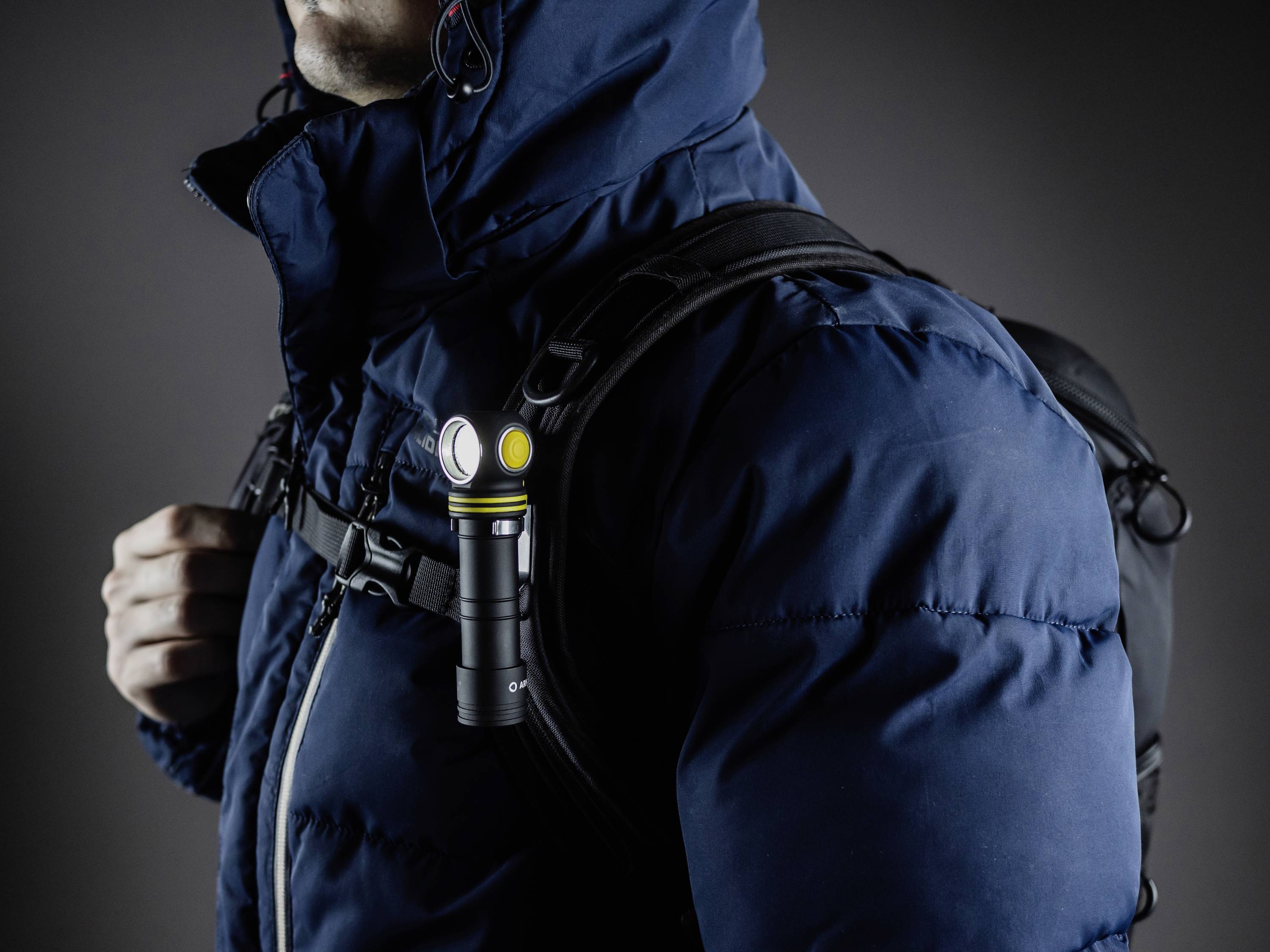A person wearing a blue winter jacket is carrying a rucksack. A small, illuminated torch is attached to one of the straps.