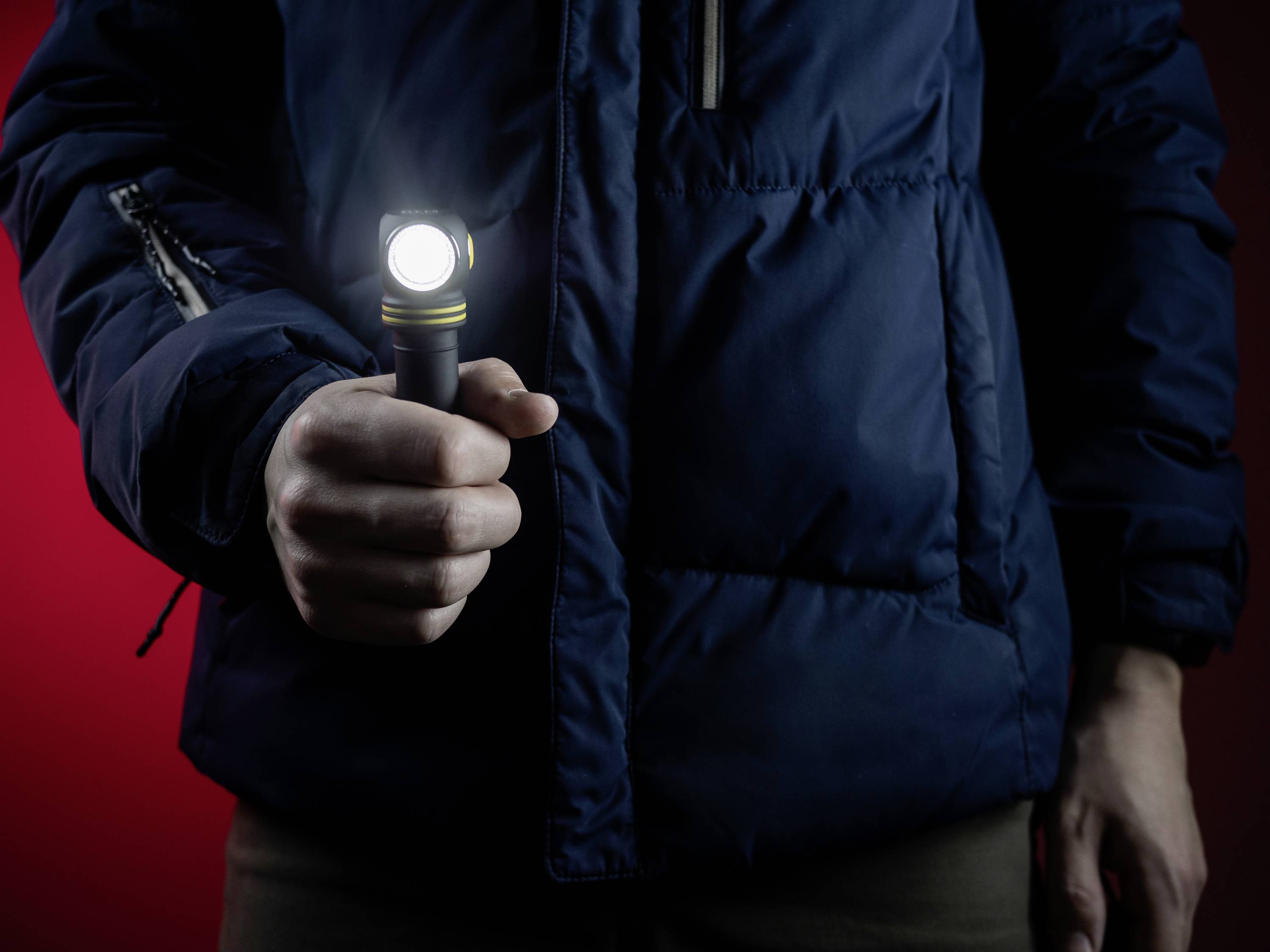 A person wearing a blue jacket is holding a switched-on torch in front of a red background.