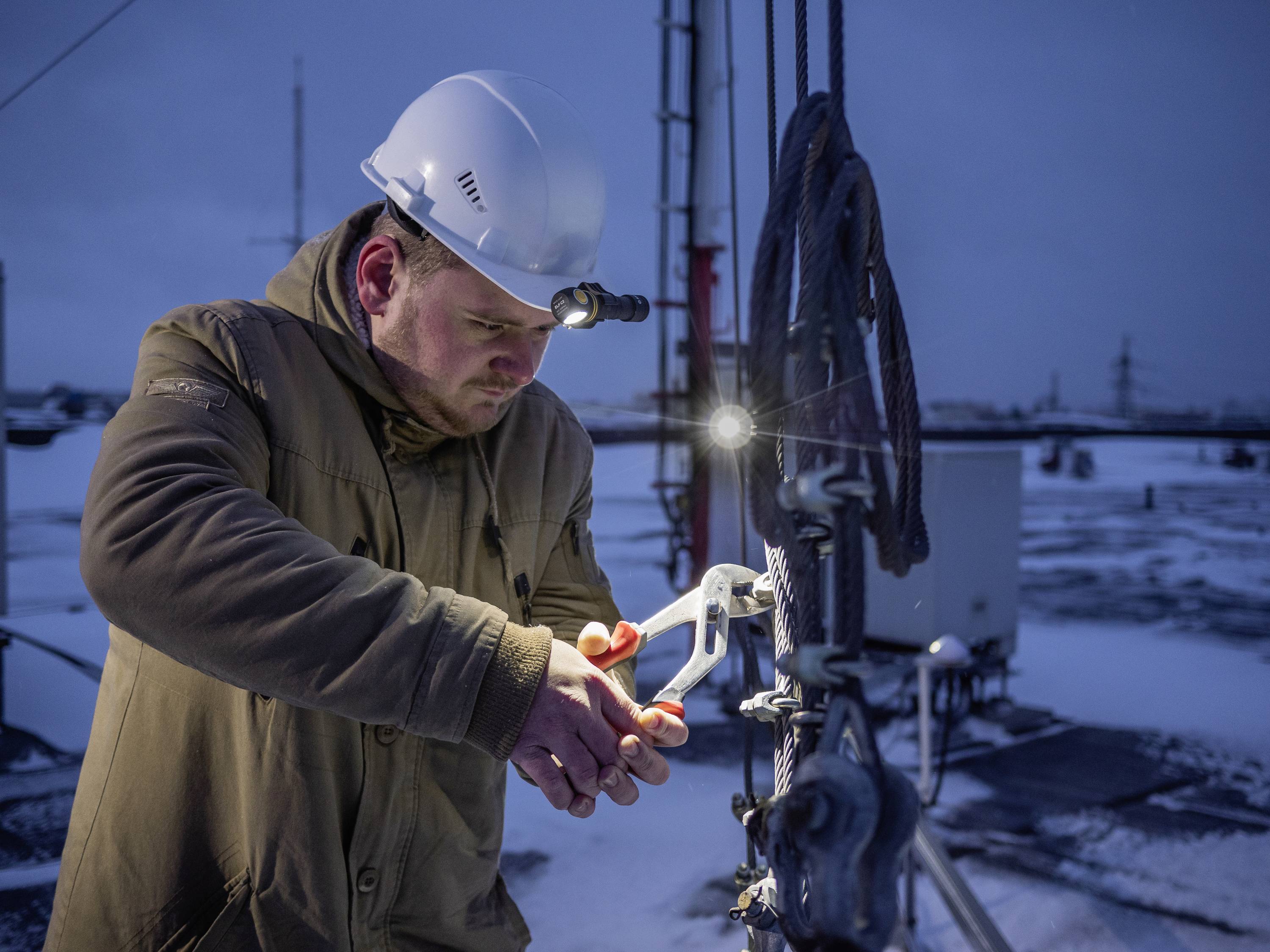 A worker in protective clothing and a helmet is repairing an installation with tools at dusk on a snow-covered industrial site.