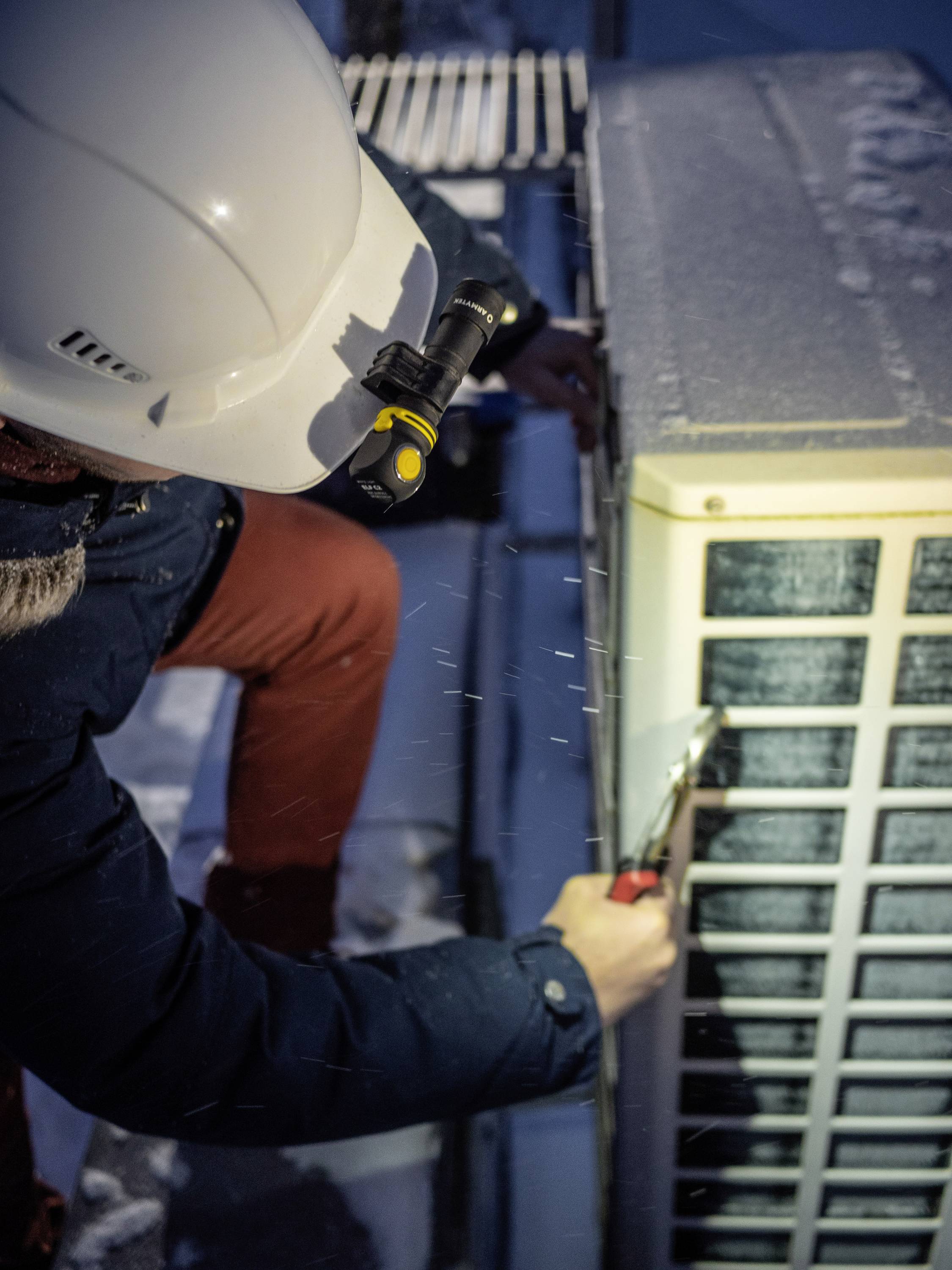 A technician is wearing a helmet and working on an air conditioning unit in the snow. He is using tools to maintain the equipment in cold conditions.