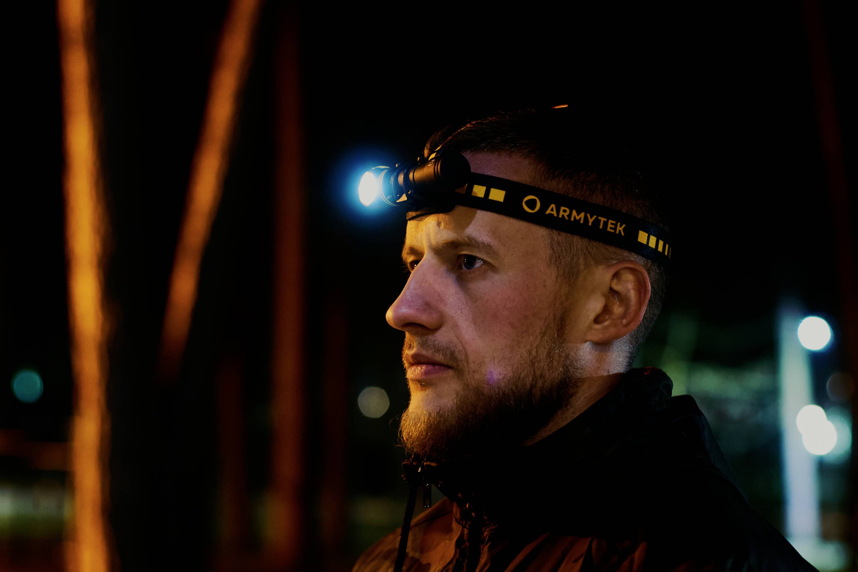 A man is wearing a head torch outdoors at night, looking intently into the distance. Trees and lights can be seen in the background.
