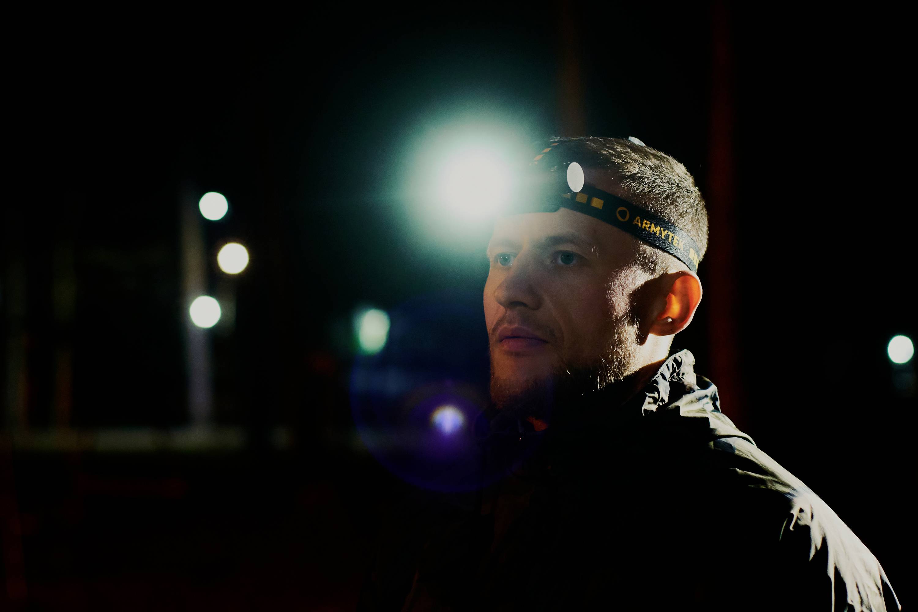 A man is wearing a headlamp in the darkness. He is outside, surrounded by blurred lights in the background.