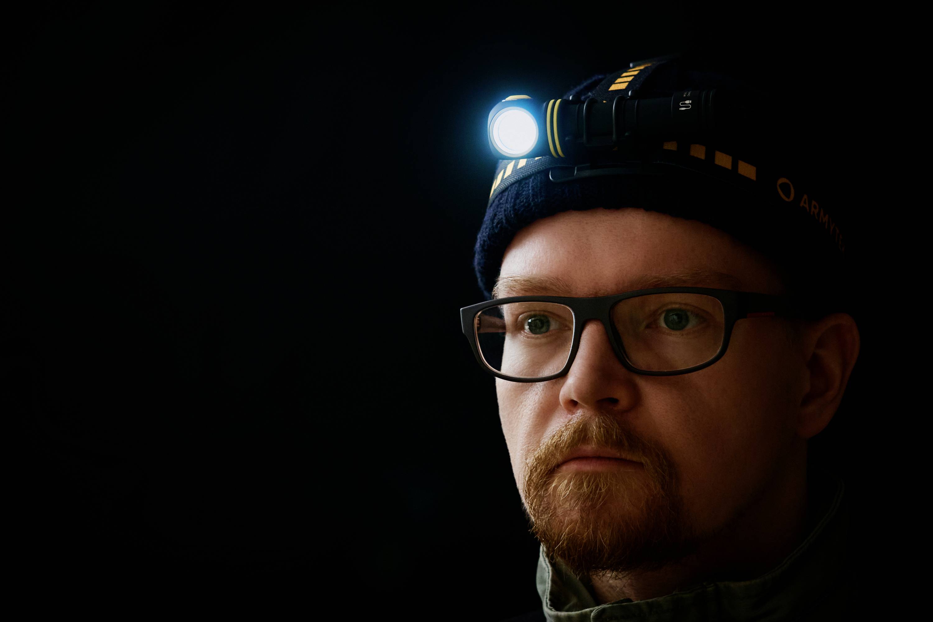 A man wearing glasses is wearing a head torch that illuminates a dark room.