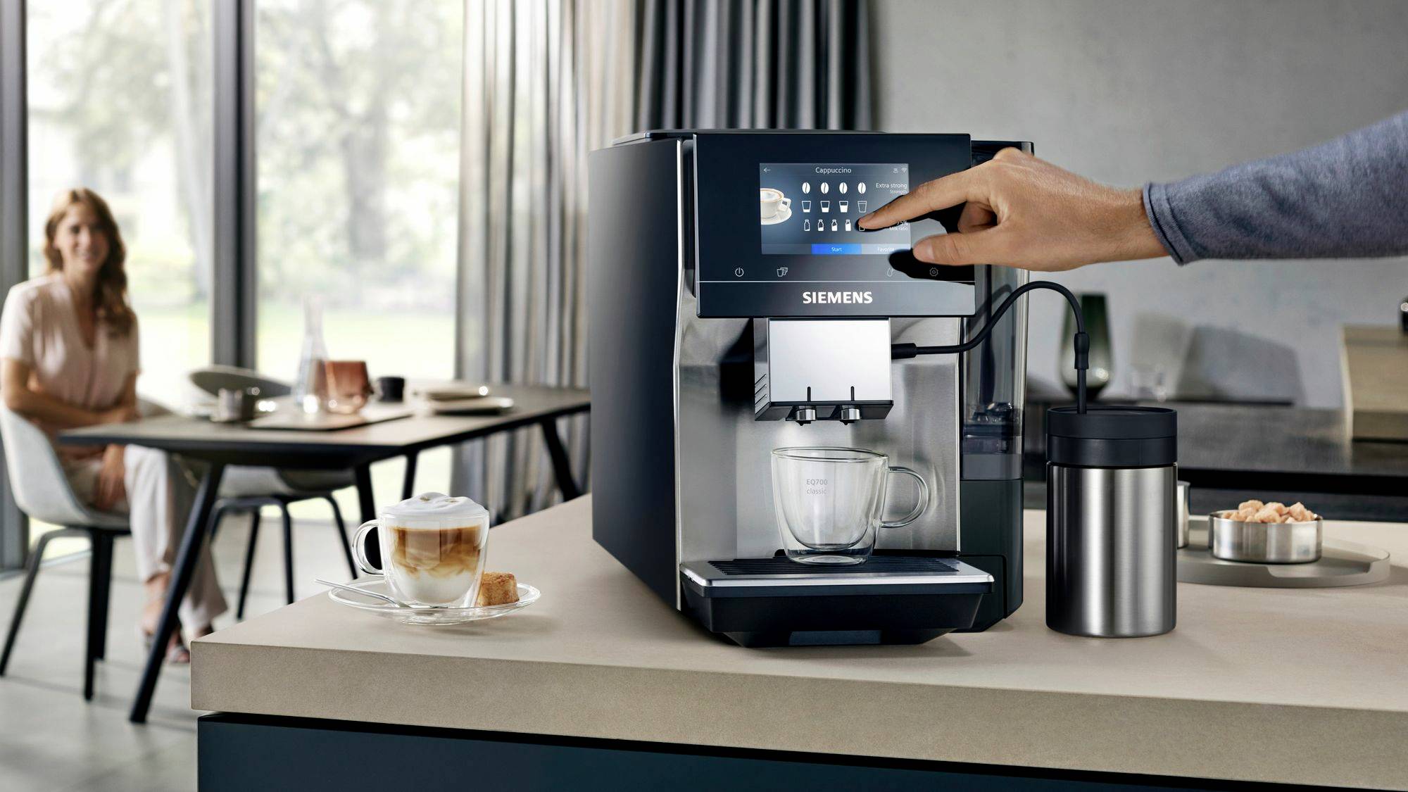 A hand operates a Siemens coffee machine on a kitchen worktop. In the background, a woman sits at the table with a cup of coffee.