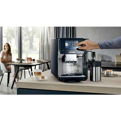 A hand operates a Siemens coffee machine on a kitchen worktop. In the background, a woman sits at the table with a cup of coffee.