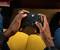 A person is sitting on a yellow sofa, holding a black video game controller with coloured buttons, ready to play.