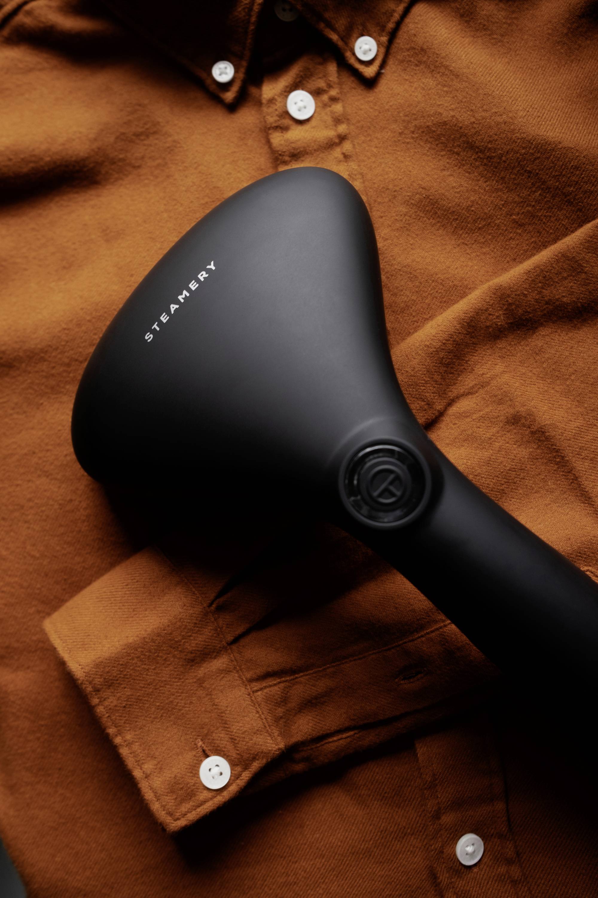 A black steam straightener from the brand STEAMERY is lying on an orange buttoned shirt.