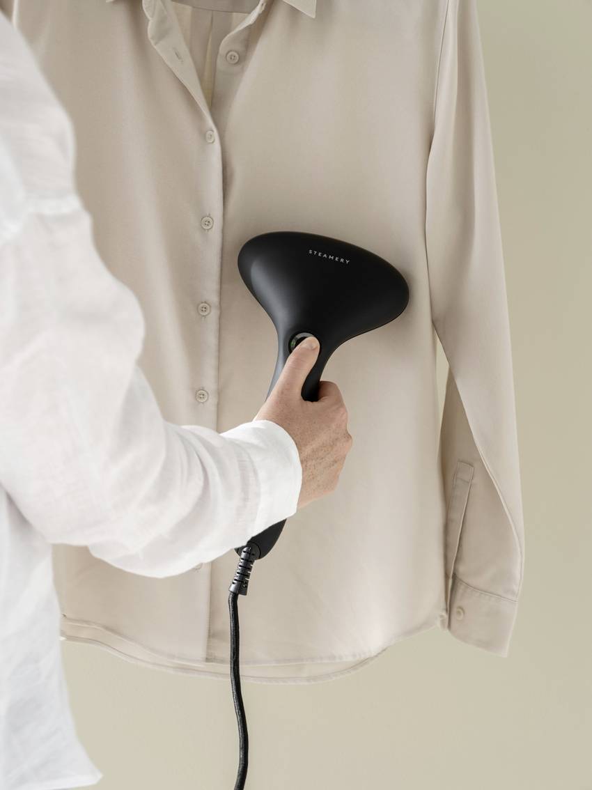 A person is steaming a light-coloured shirt with a black handheld steamer. The steamer is held in front of the shirt to remove creases.